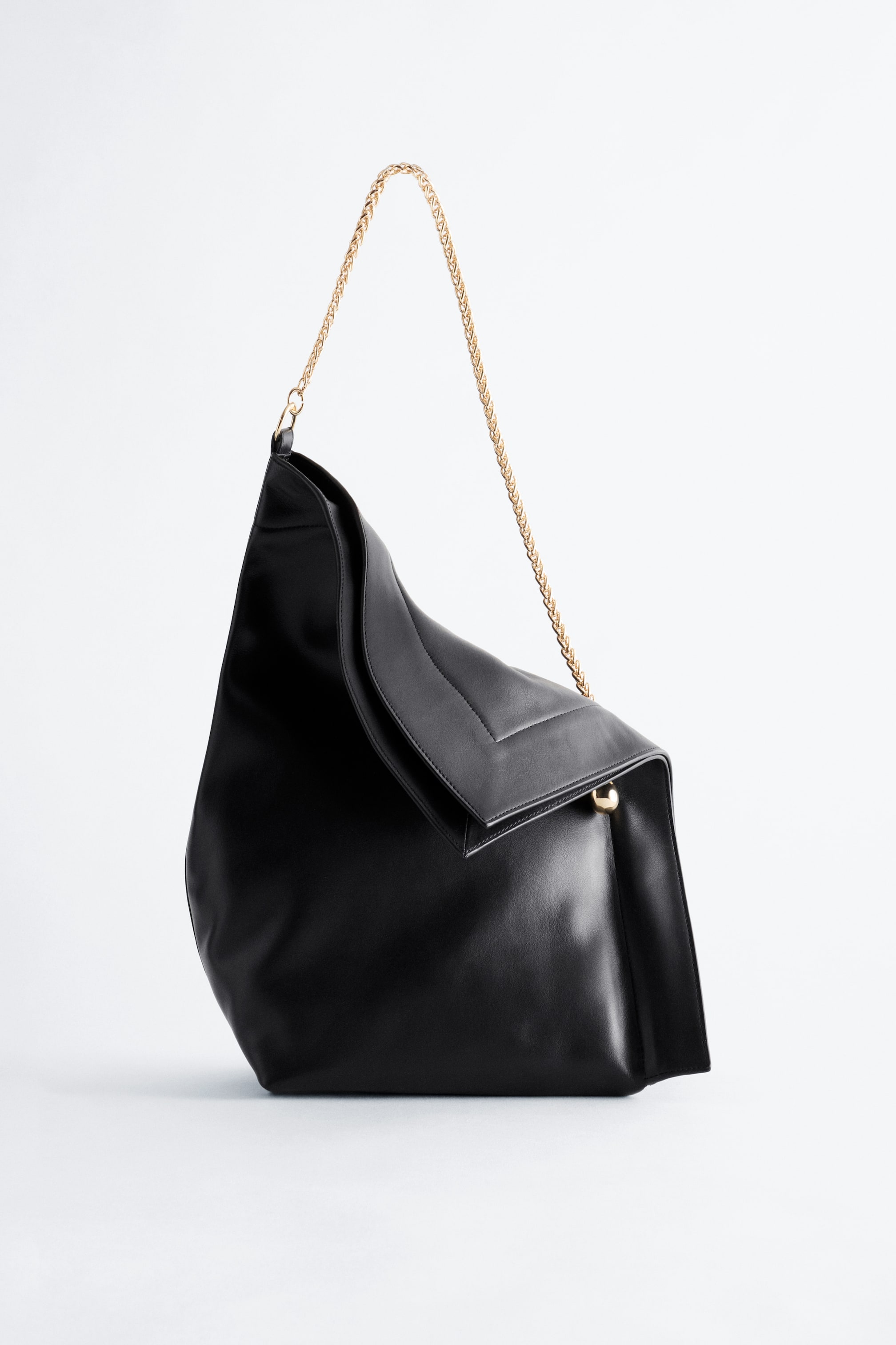 Ver imagen más grande: El bolso negro minimalista, presentado ligeramente de frente, muestra una silueta blanda y sin estructura, con una abertura superior curvada, una correa de hombro de cadena en color dorado y un diseño de solapa superpuesta en la parte superior con un pequeño botón dorado visible.