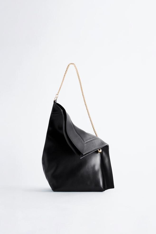 Un bolso de hombro de piel negra con una silueta suave y desestructurada está de pie, orientado hacia delante para mostrar su forma asimétrica y su abertura superior plegada. El bolso presenta una delicada correa de hombro de cadena en tono dorado y un pequeño adorno esférico en tono dorado en la esquina plegada. - 1