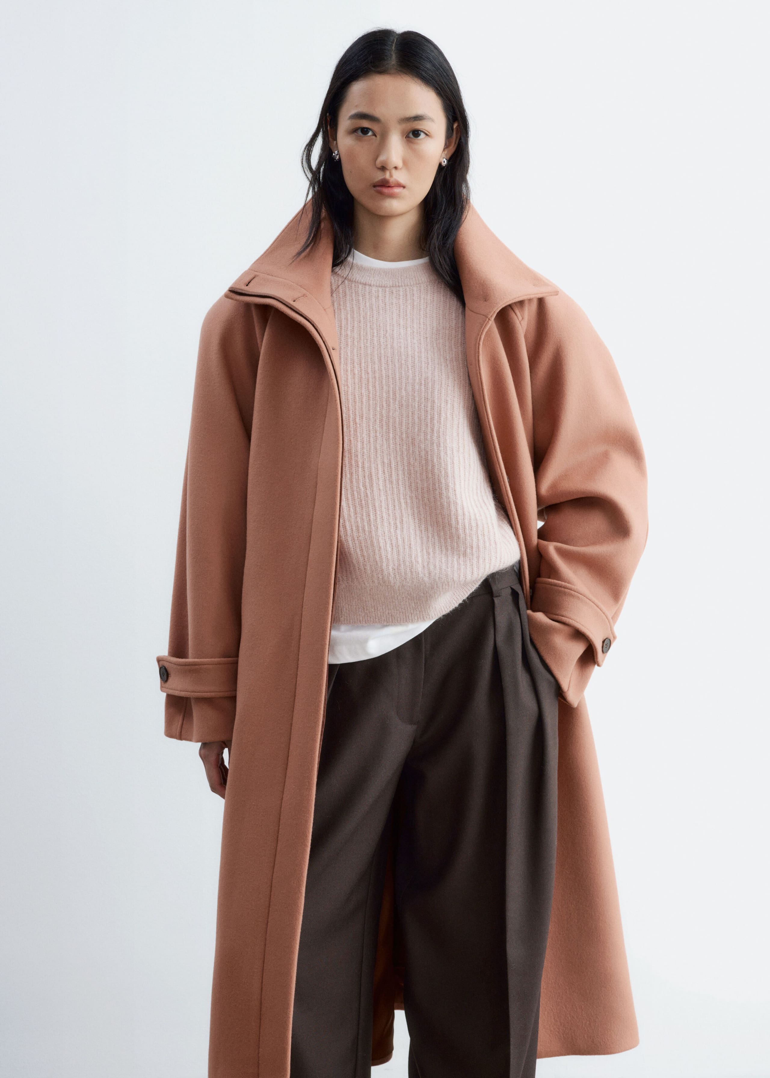 A peach-brown wool blend coat is worn open by the model, featuring a wide collar, bold shoulders, and buttoned tabs on the cuffs. The coat is layered over a light pink ribbed knit jumper and dark brown trousers.