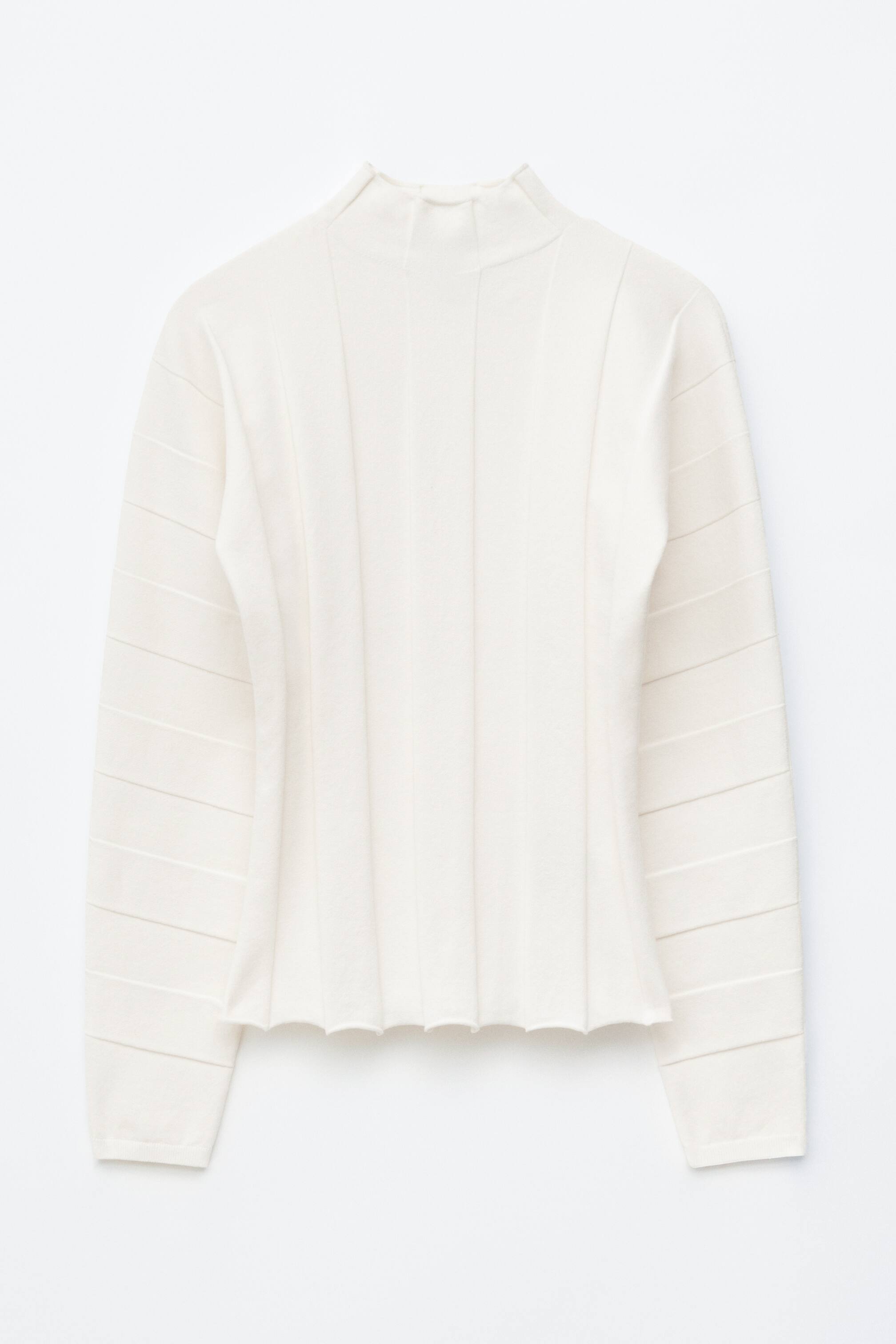 Se større billede: En cremefarvet top vises forfra med en højhalset krave. Dens hoveddel har brede, lodrette ribber, der former en tætsiddende talje, og de lange ærmer har vandrette teksturerede bånd.