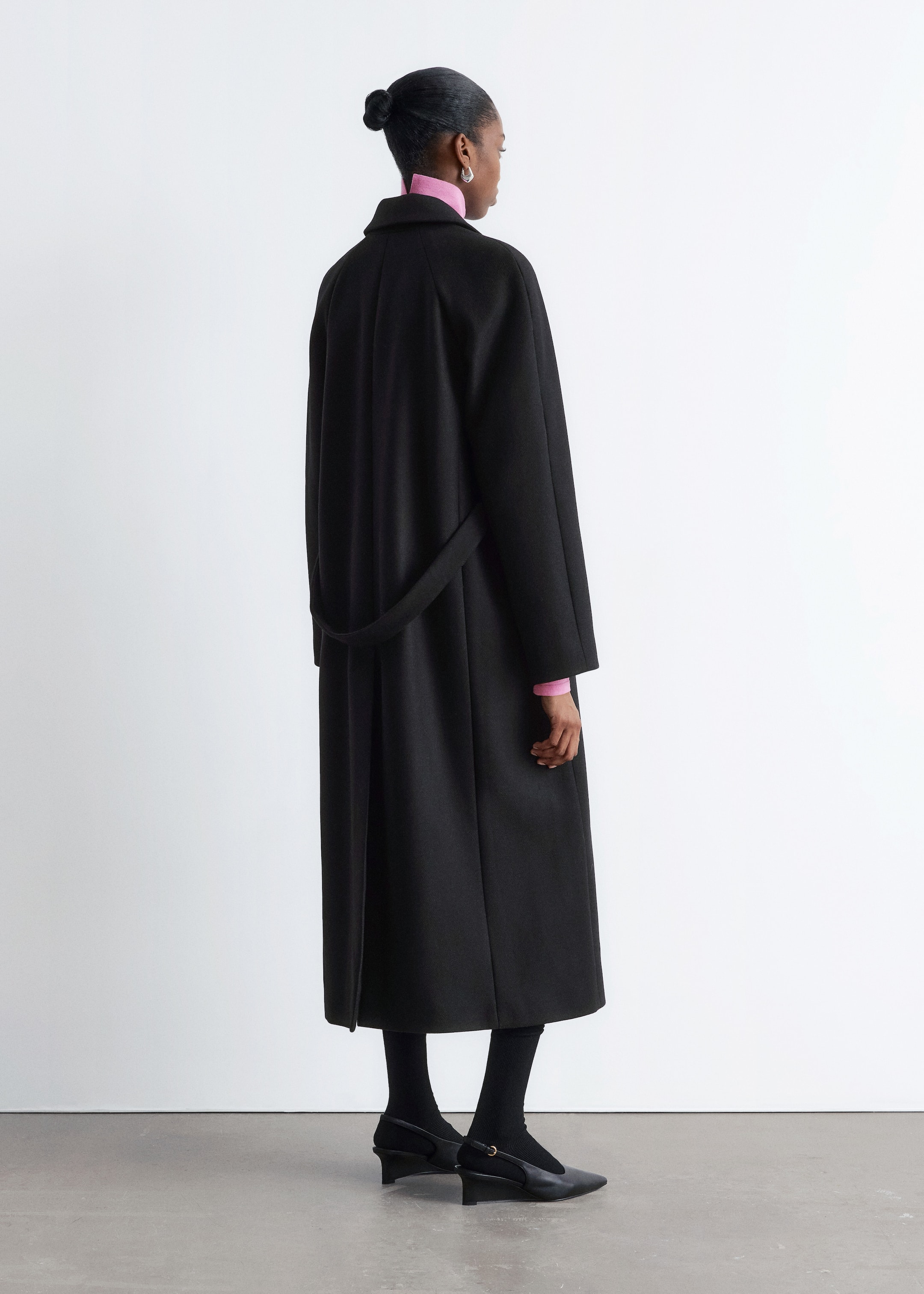 Visualizza immagine più grande: Un cappotto nero a tutta lunghezza è esposto dal retro, indossato sopra un top rosa a collo alto. Il cappotto presenta una vestibilità rilassata e dritta, maniche ampie, un colletto a fascetta e una cintura dello stesso tessuto drappeggiata sul retro.