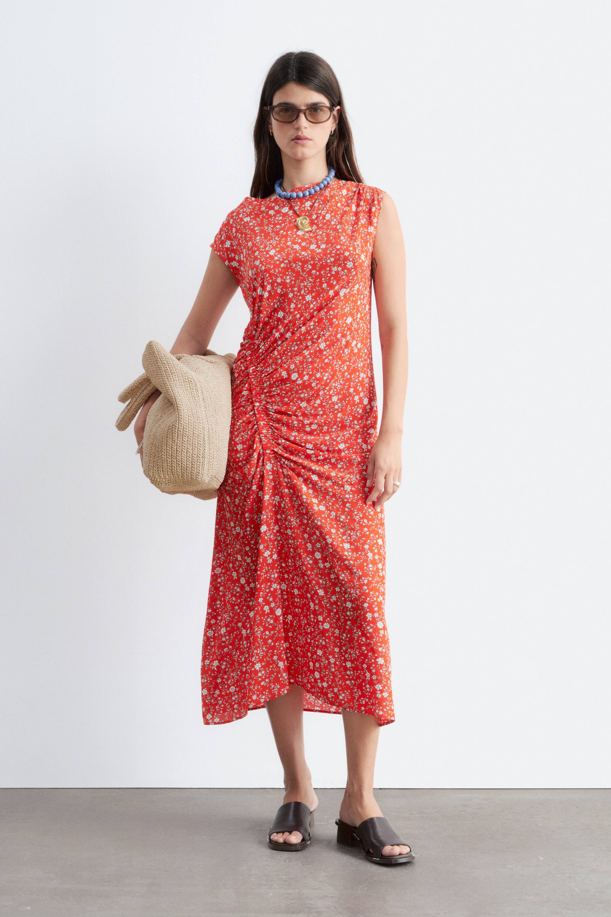 Προβολή μεγαλύτερης εικόνας: "Ένα κόκκινο midi φόρεμα με μικρό λευκό λουλουδάτο μοτίβο και κοντά μανίκια καπ. Το φόρεμα διαθέτει πτυχώσεις κατά μήκος της μιας πλευράς, δημιουργώντας μια υφή με συγκεντρωμένες πτυχές."