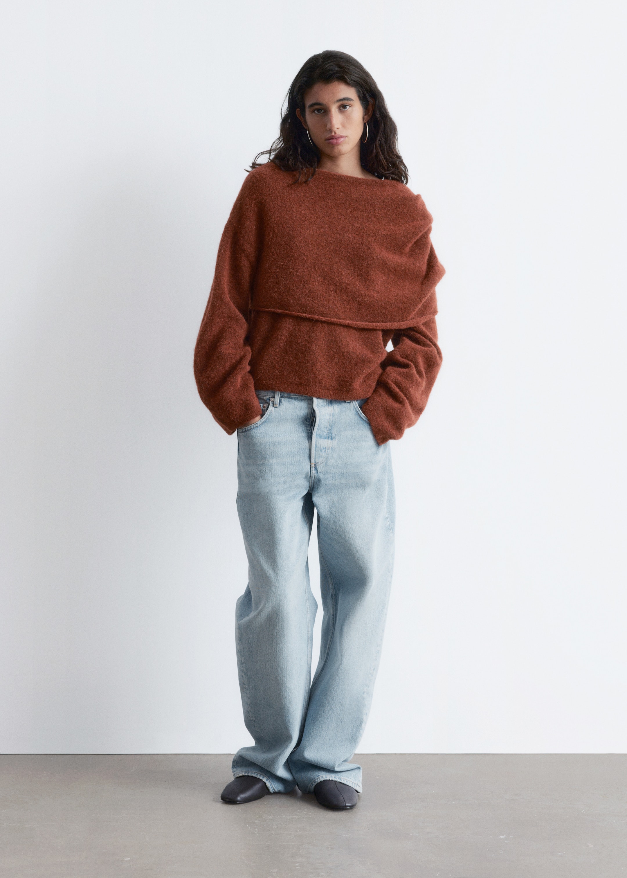Se større billede: En rustfarvet, fuzzy strikket sweater præsenterer en bred, draperet halsudskæring og lange, løse ærmer, der vender fremad for at fremvise dens afslappede pasform. Den er stylet med lyse, højtaljede, bredbenede denimjeans, der samler sig over mørke, flade, lukkede sko.