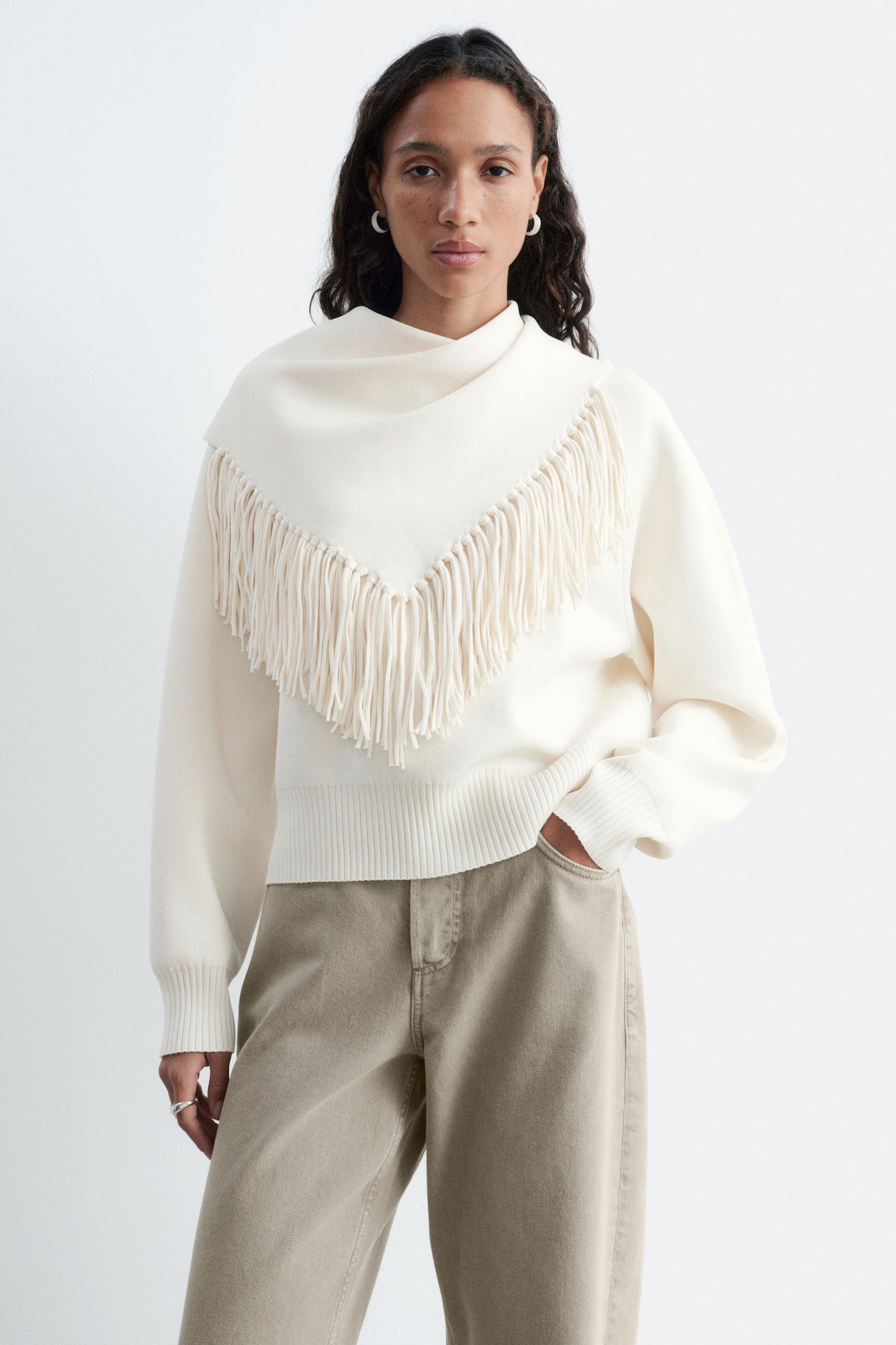 Visualizza immagine più grande: Un maglione lavorato a maglia color crema, caratterizzato da un dettaglio di sciarpa integrata con drappeggio e da una lunga frangia a V, indossato dalla modella. Il maglione presenta maniche lunghe con polsini a coste e orlo a coste, ed è abbinato a pantaloni color oliva chiaro.