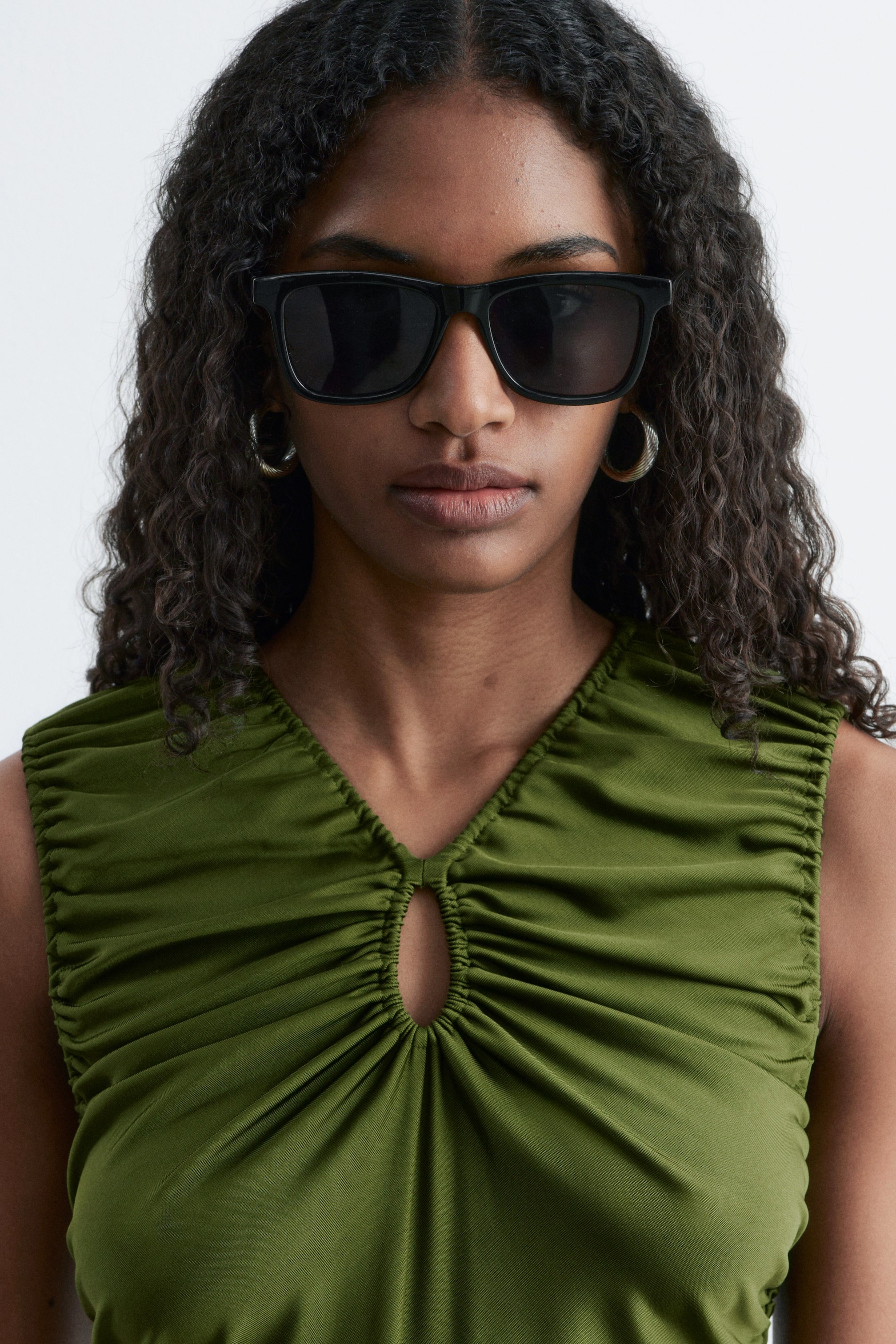 Nagyobb kép megtekintése: Egy zöld, ujjatlan felső V-nyakkivágással és középső kulcslyuk kivágással rendelkezik. A felső összegyűrt anyagot mutat be, amely ráncolt hatást kelt a nyakkivágás mentén és az elején lefelé.