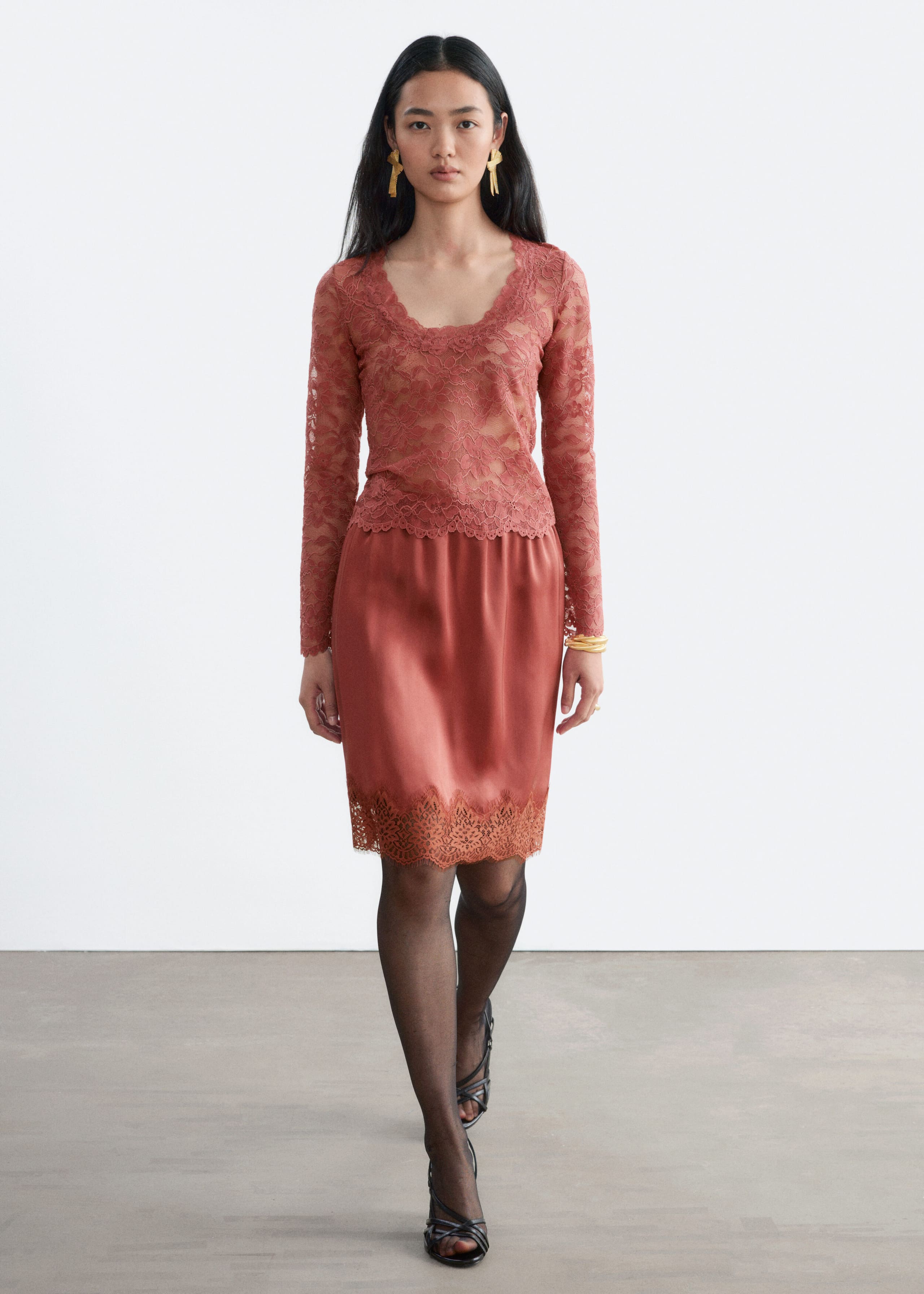 A long-sleeved, rust-coloured lace top featuring a scoop neckline and scalloped edges is being worn by the model, paired with a matching satin knee-length skirt with a wide lace-trimmed hem. The outfit is styled with sheer black tights and strappy black heels as the model walks forward.