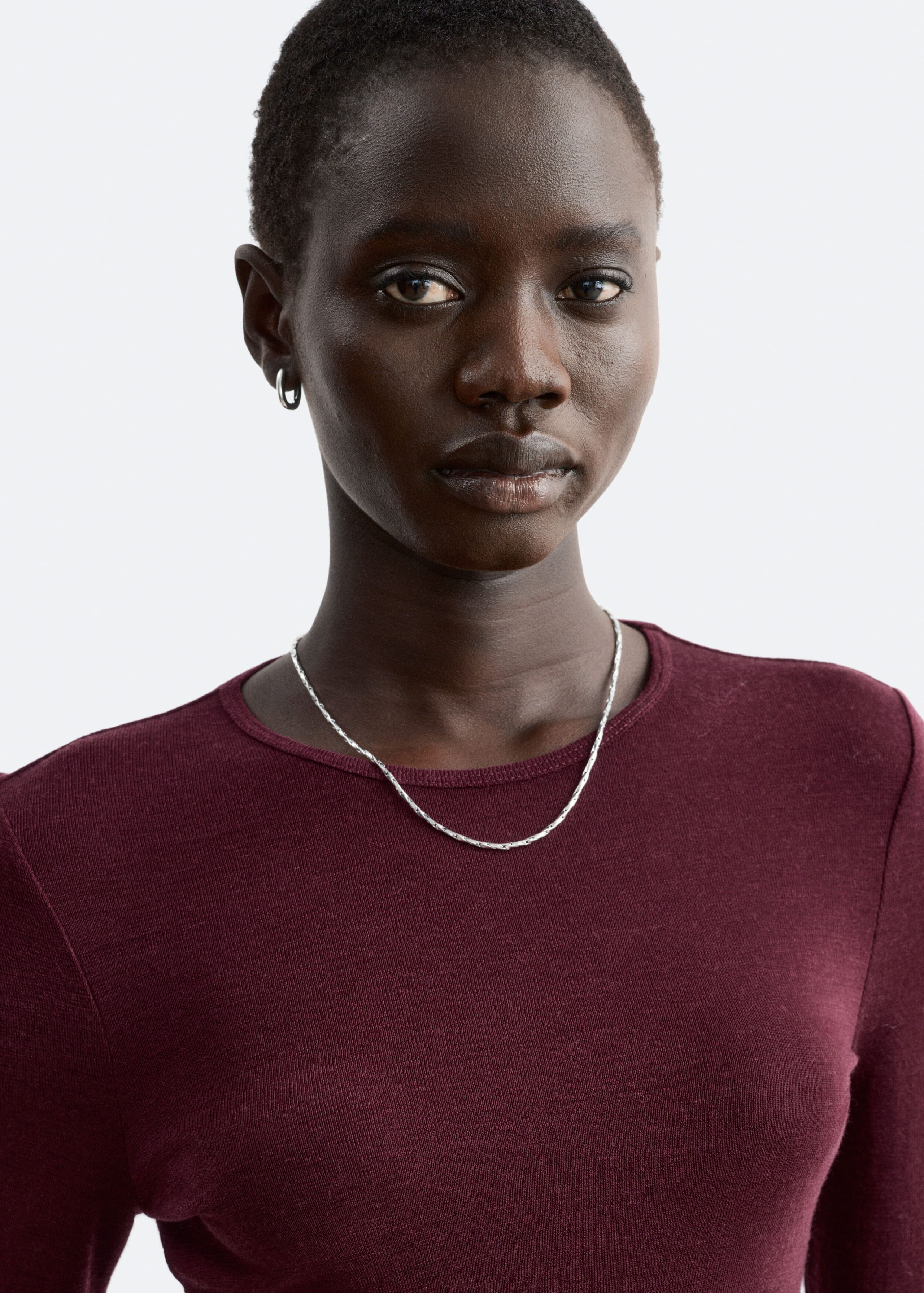 Ein schlichter Wollpullover in tiefem Burgunderrot wird vom Model getragen. Das Oberteil verfügt über einen Rundhalsausschnitt und eine feine Strickstruktur und ist mit einer dünnen silbernen Kette kombiniert, die am Kragen anliegt.