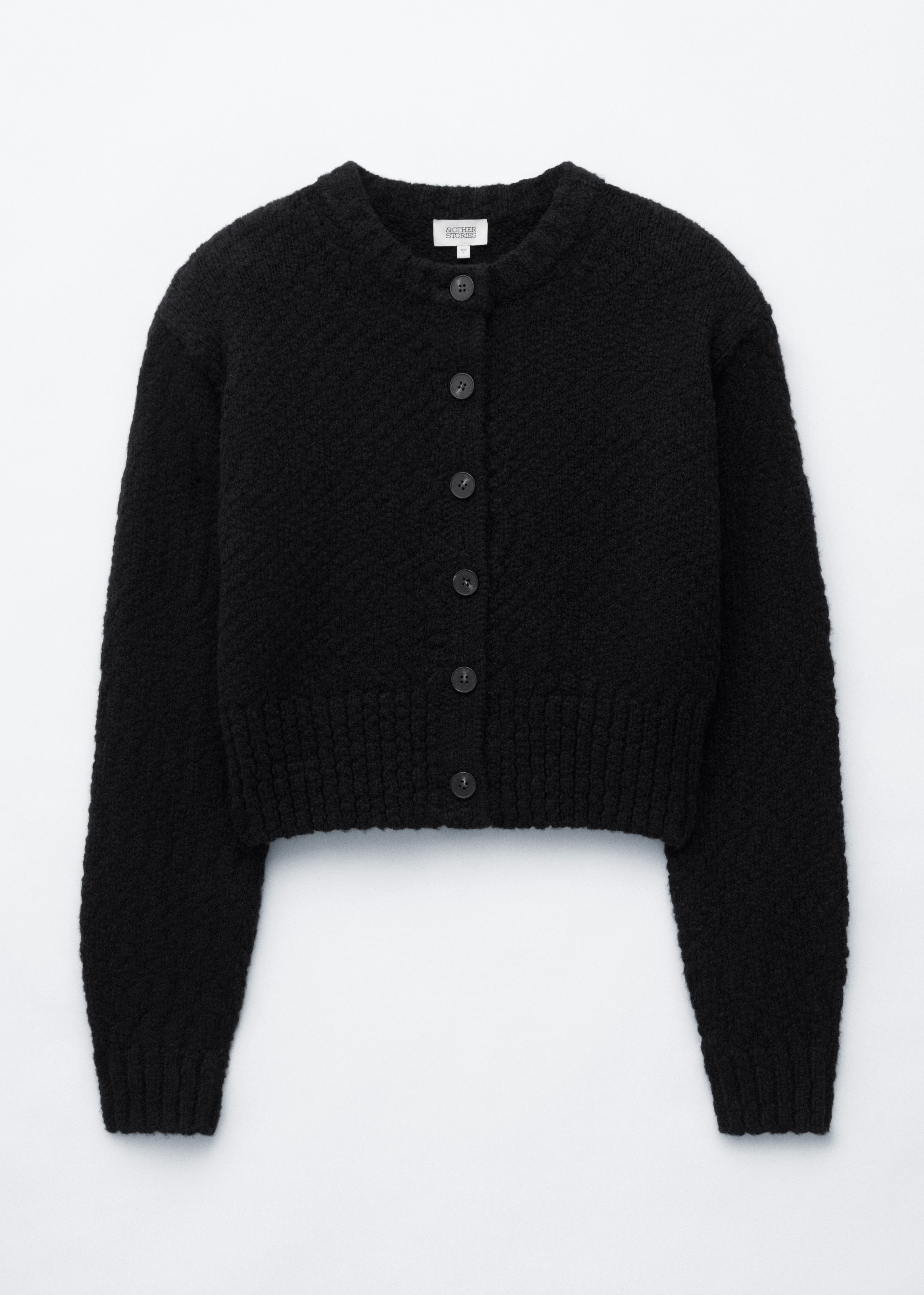 A black wool-cotton cardigan featuring a textured knit and front button fastening is worn open by the model. The cardigan is layered over a blue-and-white striped shirt and cream trousers, while a patterned woven handbag is being held.