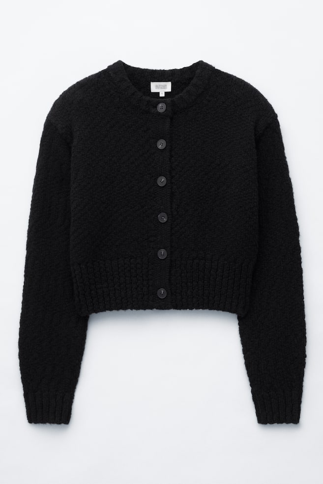 Un cardigan nero in lana e cotone è mostrato frontalmente, caratterizzato da una lavorazione a maglia testurizzata, girocollo e chiusura frontale con sei bottoni scuri. Il modello cropped mette in risalto le maniche lunghe con polsini a coste e un ampio bordo a coste. - 1