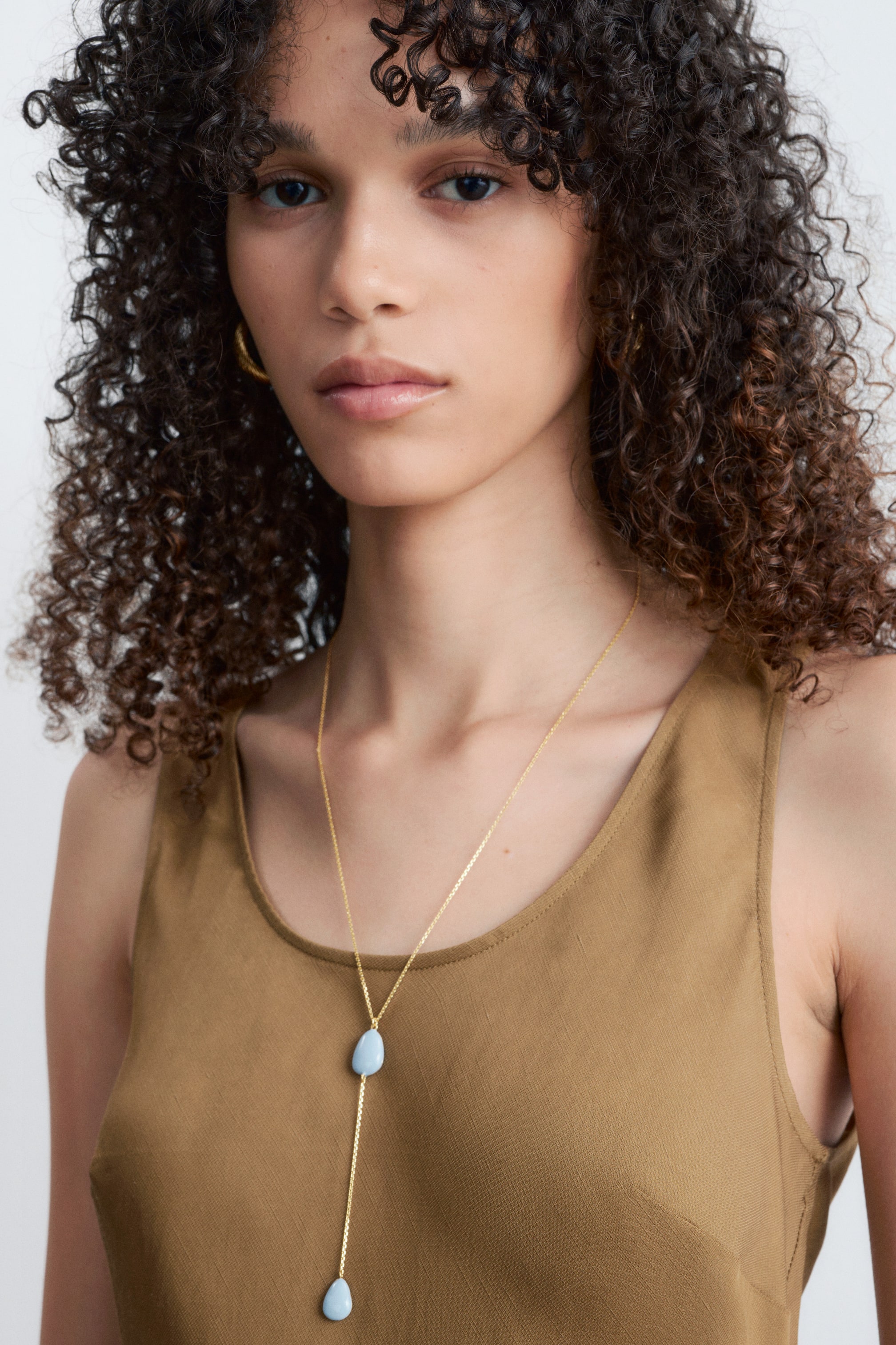 Größeres Bild ansehen: Eine goldfarbene Lariat-Halskette mit zwei unregelmäßig geformten, hellblauen Steinen. Ein Stein ist höher an der Kette positioniert, während der zweite Stein am Ende eines längeren Kettenabschnitts baumelt und auf einem beigen Oberteil ruht.