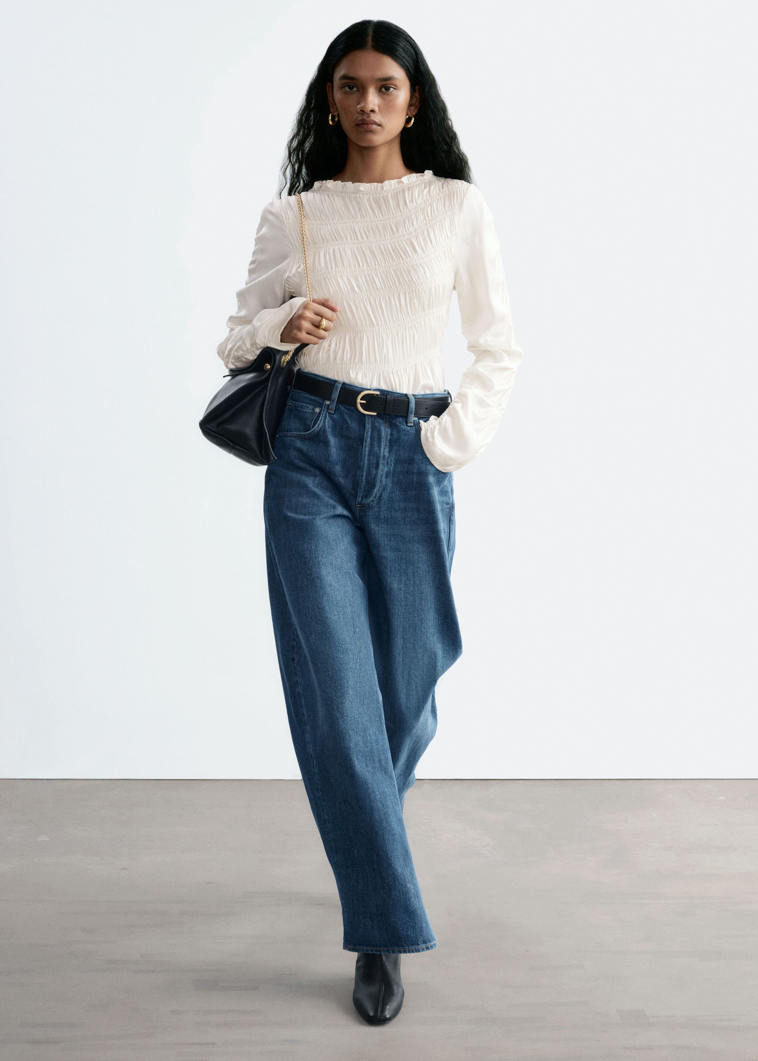 Medium blue denim jeans with an oversized barrel-leg silhouette are being worn by the model. The jeans are styled with a black leather belt, a tucked-in white smocked blouse, and black leather boots.