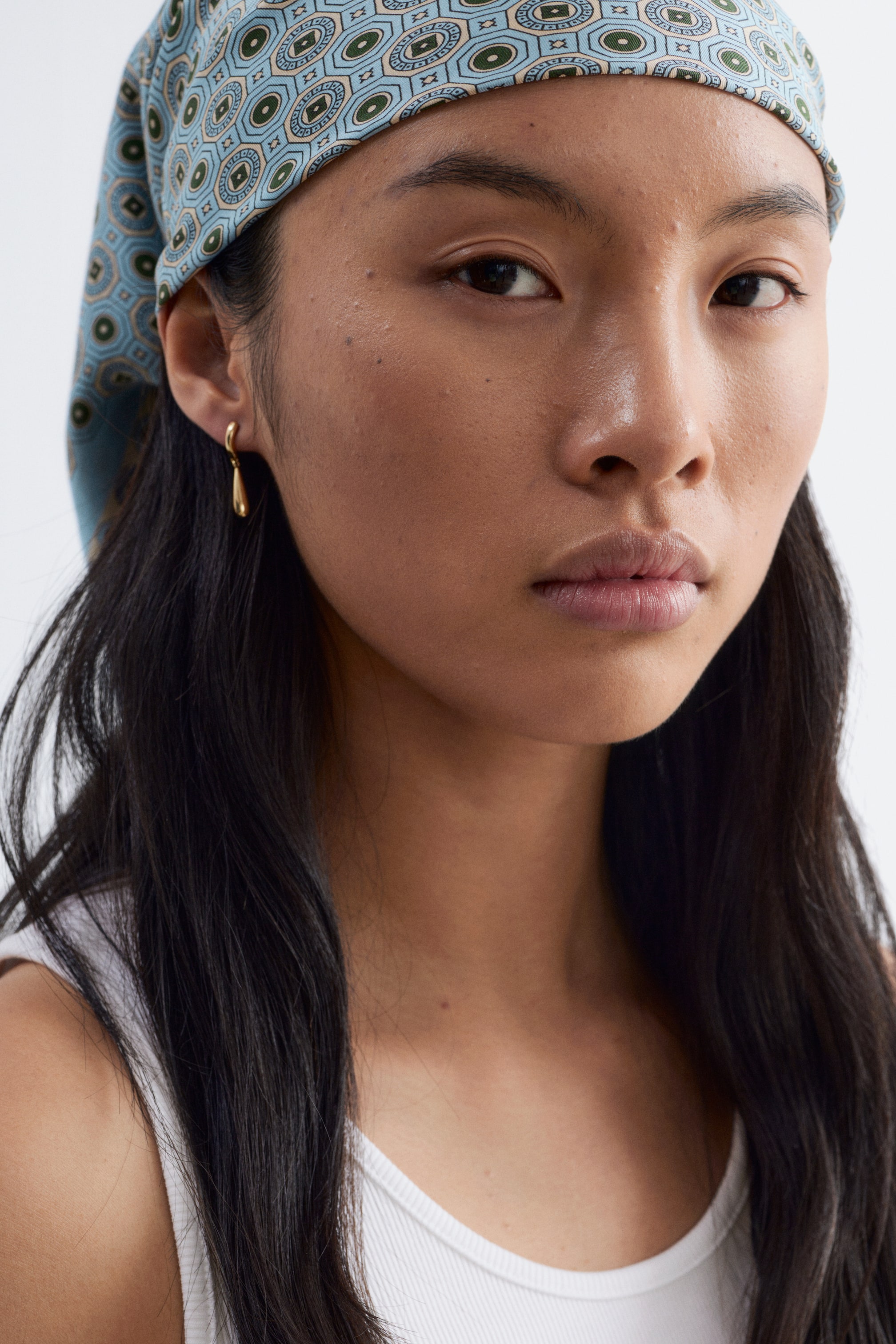 Agrandir l'image: Un bandana en tissu bleu clair est porté sur la tête, arborant un motif géométrique composé de motifs octogonaux et circulaires de couleur bleu foncé, vert et beige. Une boucle d'oreille en forme de goutte d'or pend du lobe de l'oreille, visible du côté droit.