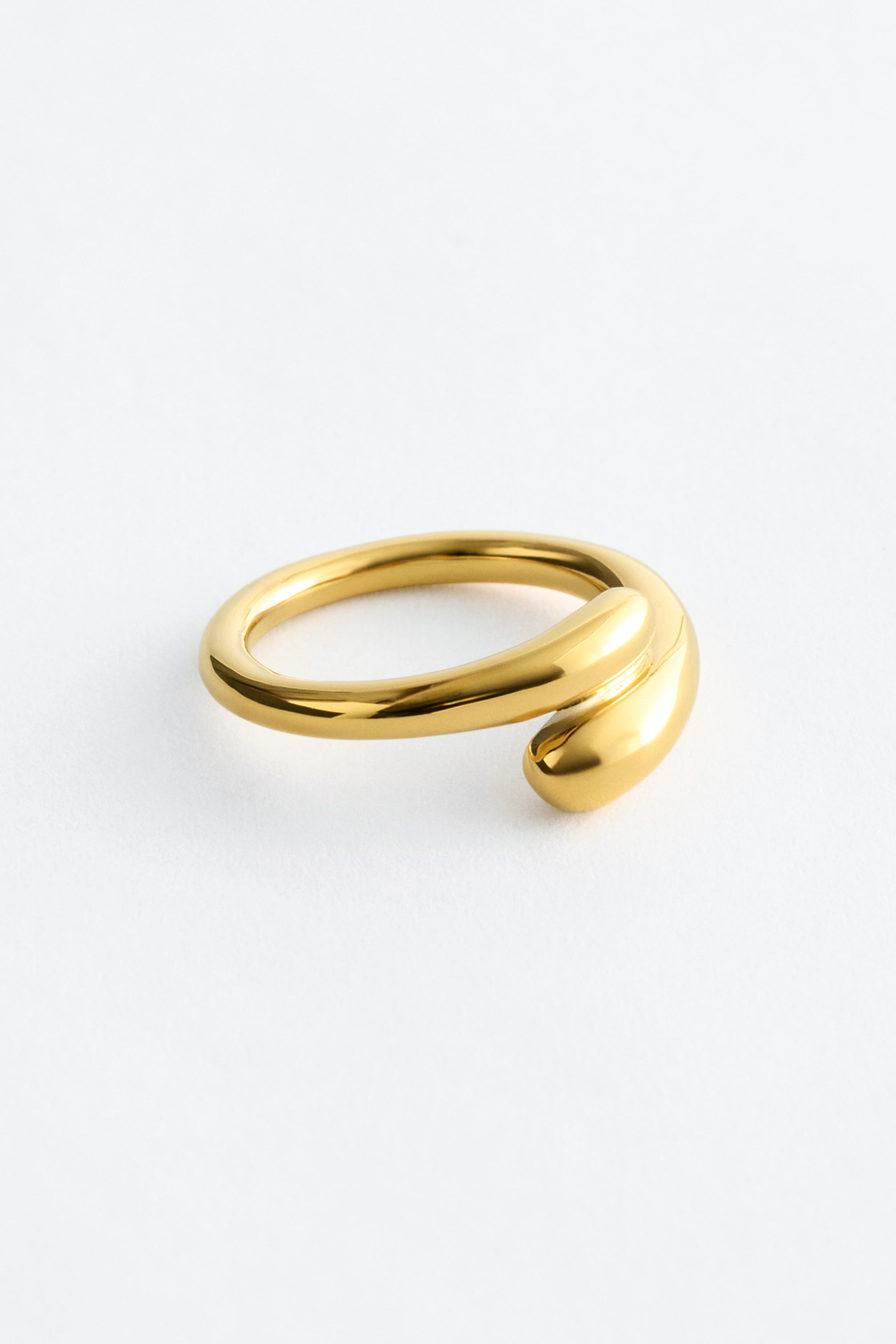 Größeres Bild ansehen: Der goldfarbene offene Ring zeichnet sich durch ein glattes, abgerundetes Band aus. Ein Ende des Bandes bildet eine abgerundete Tropfenform, während das andere Ende sich um das erste wickelt und es überlappt.
