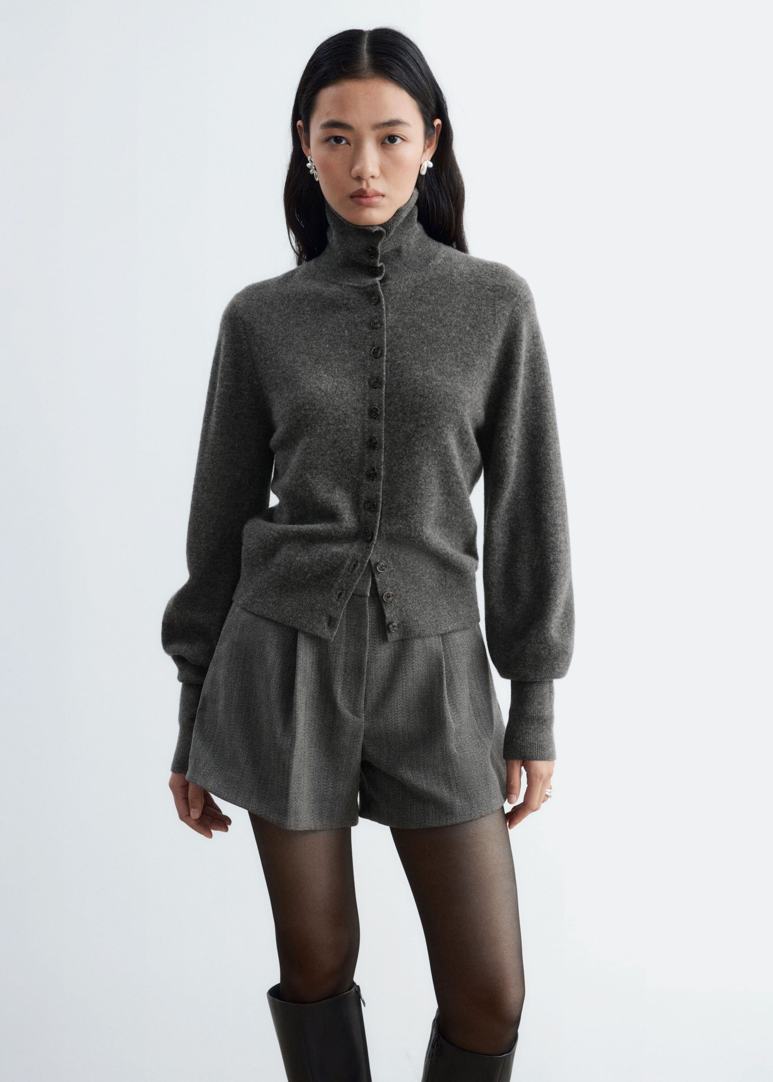 These grey, high-waisted wool-blend shorts with front pleats and an A-line silhouette are worn by the model. They are styled with a matching grey button-up turtleneck cardigan, sheer black tights, and knee-high black boots.