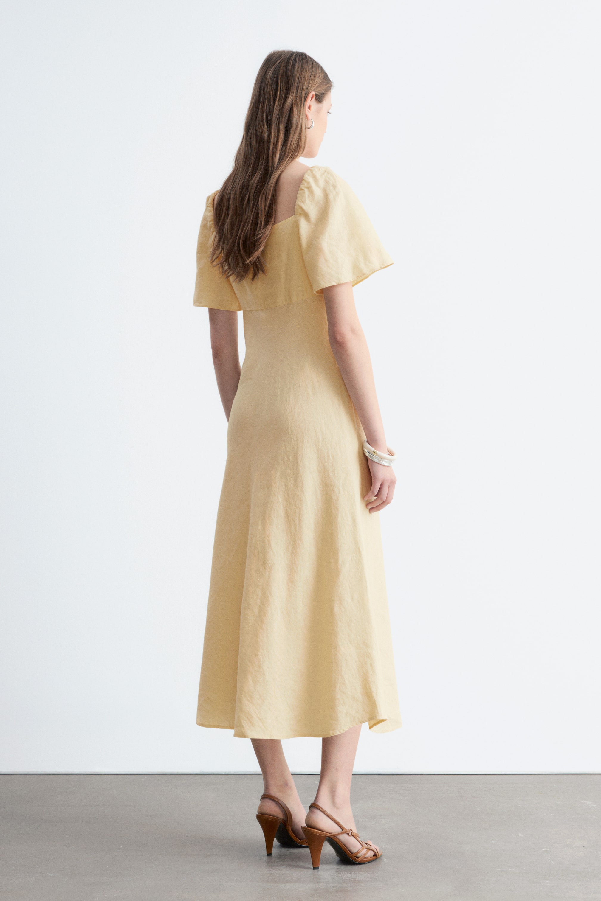 Agrandir l'image: La modèle porte une robe midi en lin jaune clair, se tenant de dos pour mettre en valeur ses manches courtes à volants, son dos droit et sa jupe évasée. La tenue est complétée par des sandales à brides marron et un bracelet en argent.