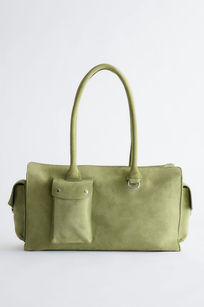 Se muestra un bolso tote de ante verde oliva desde el frontal, con una silueta estructurada y angular y dos finas asas de hombro. El diseño frontal incluye un bolsillo vertical tipo sobre con botones de presión metálicos plateados a la izquierda, un pequeño detalle de anilla en D metálica plateada a la derecha y distintivos paneles laterales con solapa. - 1