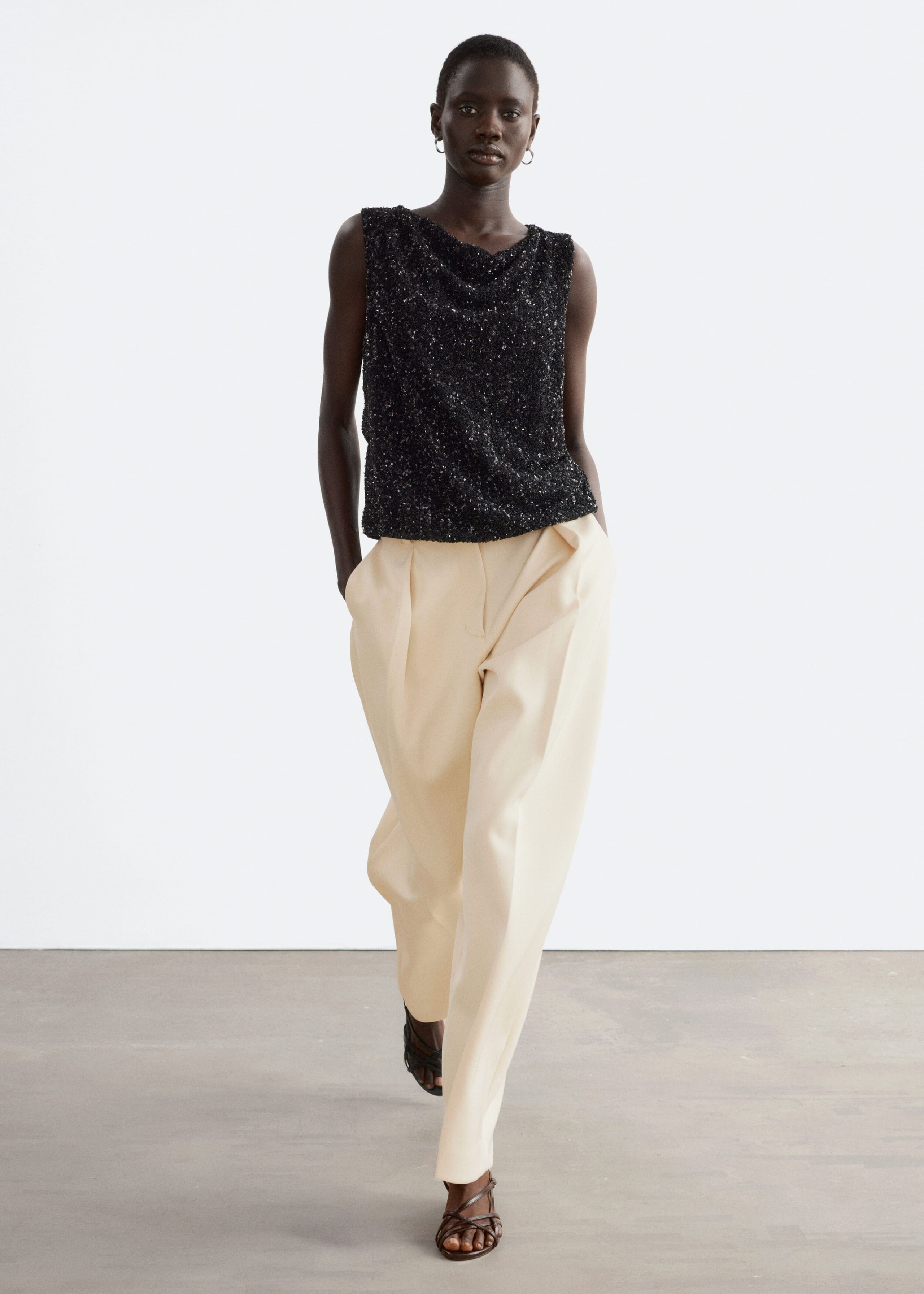 A black sleeveless top covered in sequins and featuring a draped neckline is being worn by the model. The top is paired with cream-coloured, wide-leg trousers as the model is walking forwards.