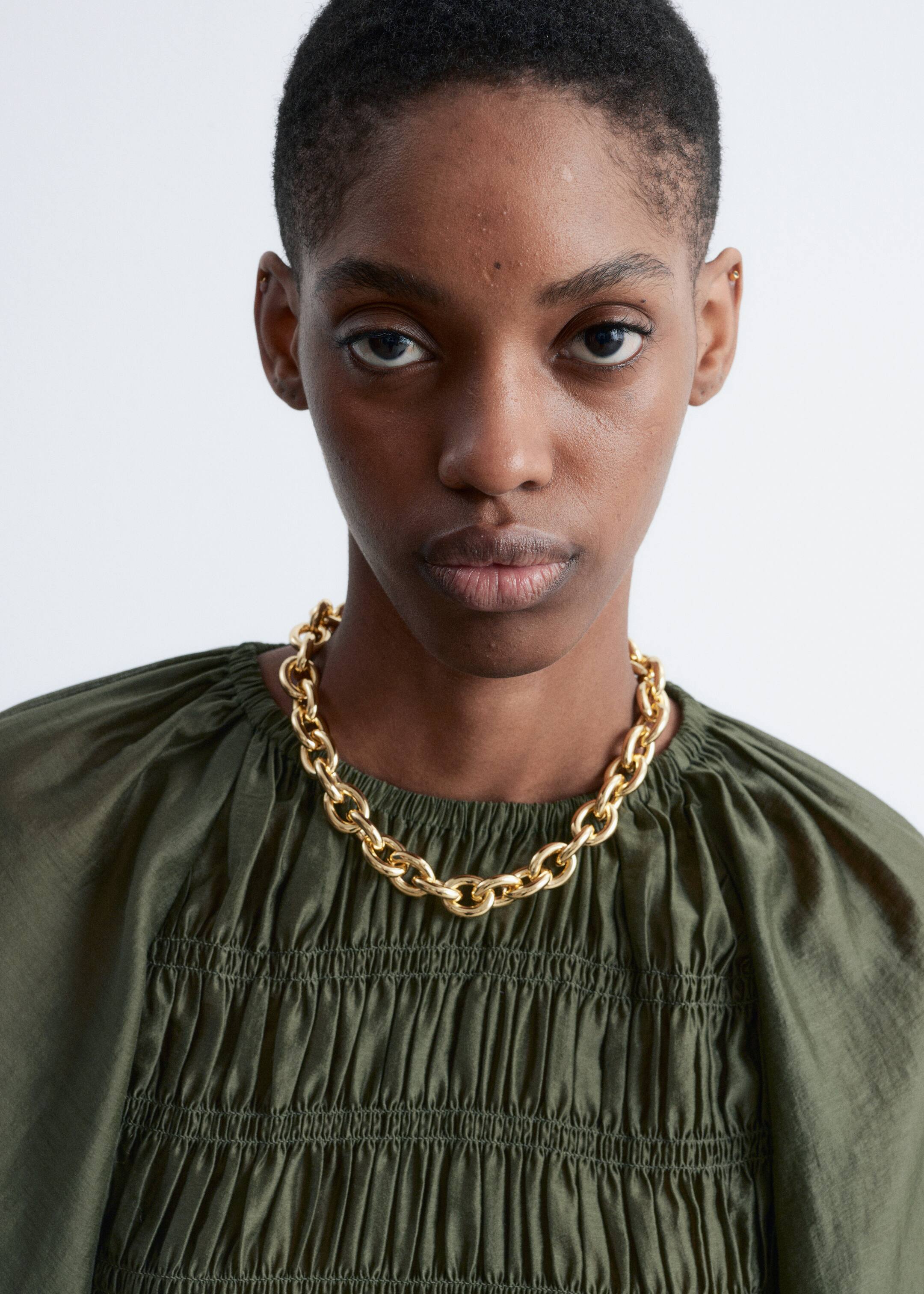 View larger image: A chunky, gold-toned chain necklace is resting on a dark olive green garment. Large, interlocked oval links are forming the necklace, which is lying flat against the gathered fabric of the top.