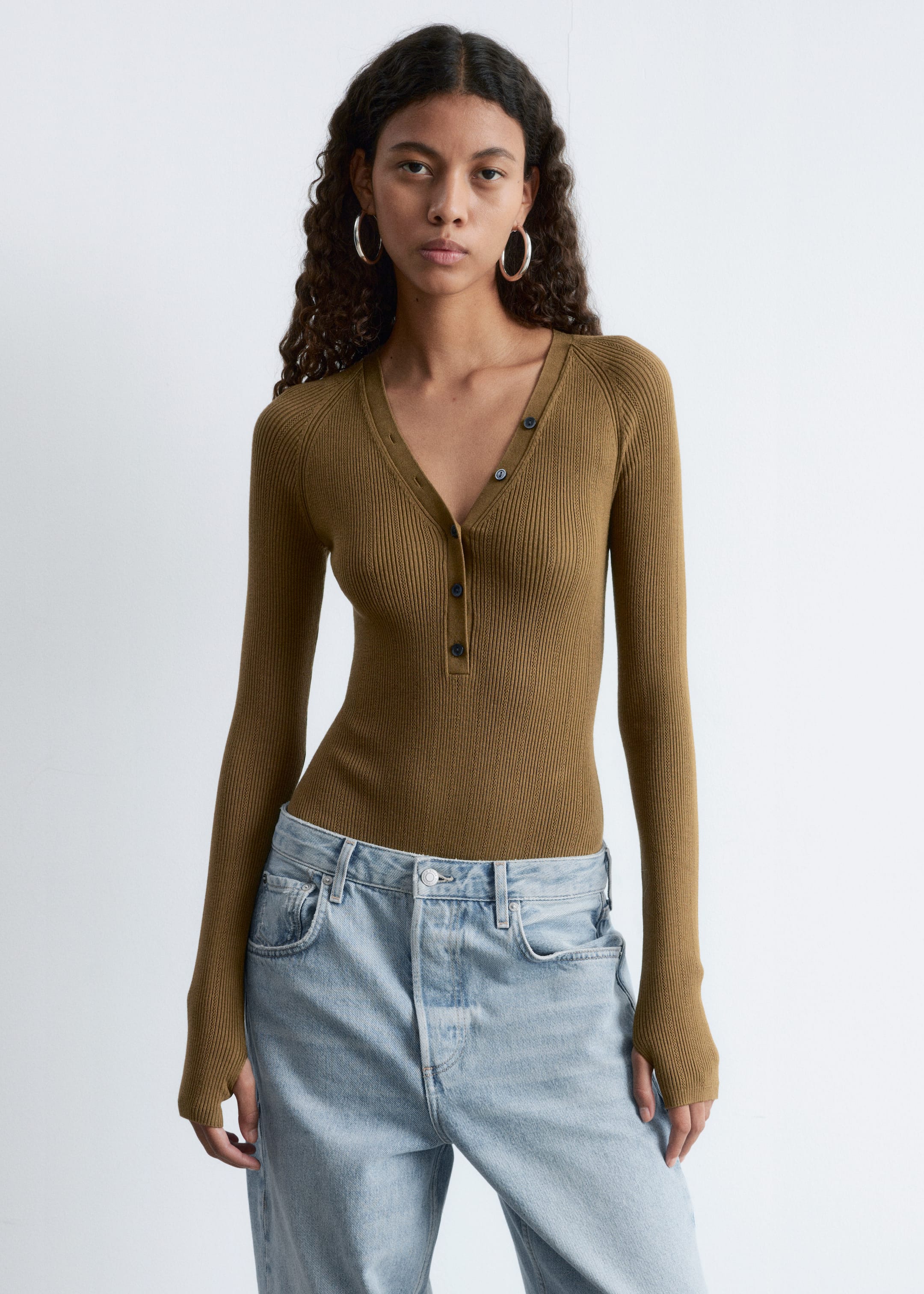 View larger image: An olive green rib-knit bodysuit with a V-neckline and partially unbuttoned front closure is being worn by the model. It features long, raglan-cut sleeves with thumbhole openings and is tucked into light wash jeans.
