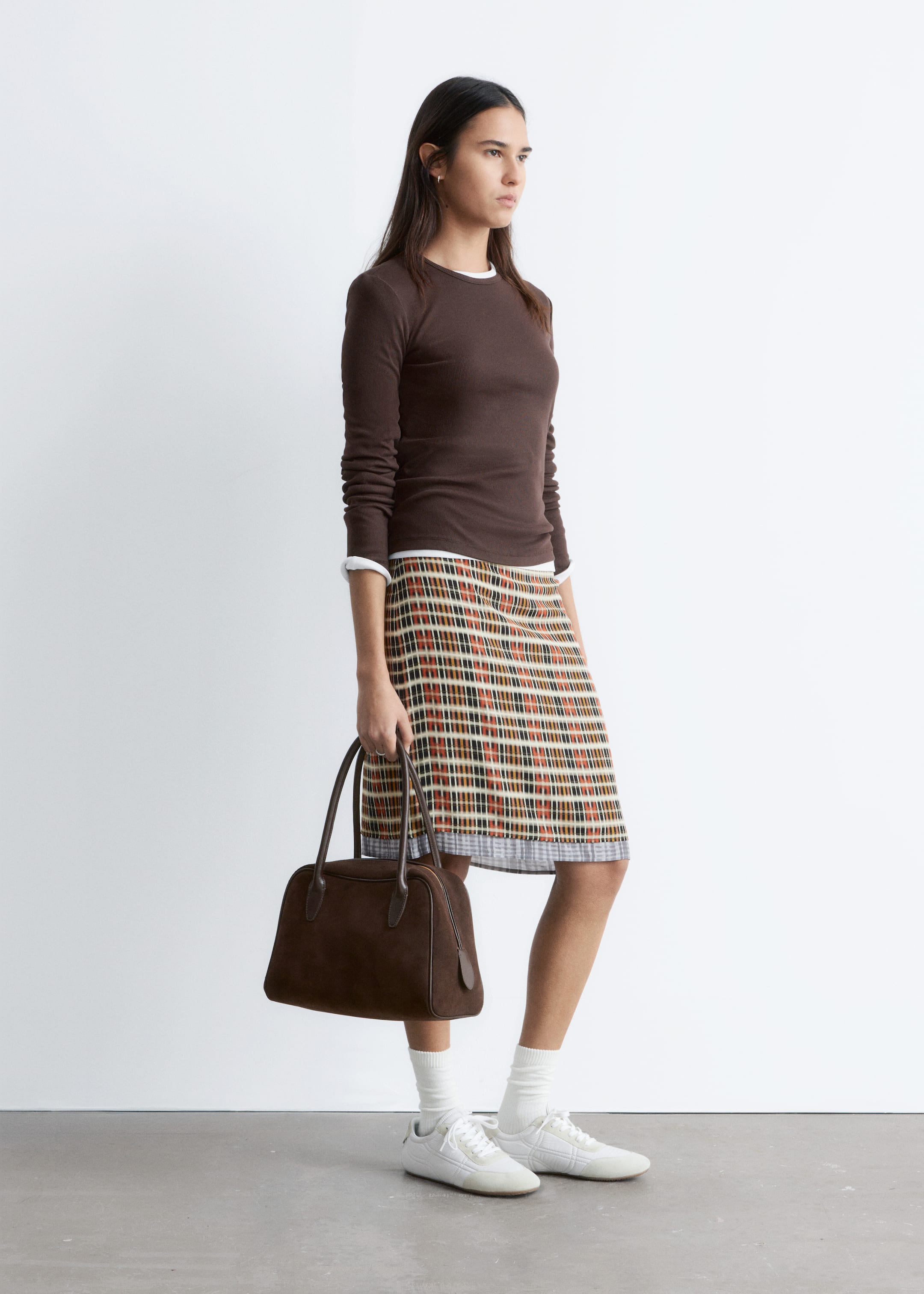 Nagyobb kép megtekintése: Egy barna hosszú ujjú felső van rétegezve egy látható fehér kerek nyakú felső fölé, amelyet egy A-vonalú mini szoknyával párosítanak, amely többszínű kockás mintázatot mutat barnában, narancsban, feketében és fehérben, a szegélynél kilátszó fehér béléssel. Egy szerkezetes barna kézitáskát tartanak, amelynek két felső fogantyúja van, és fehér sportcipőt viselnek, ráncolt fehér bokazoknival.