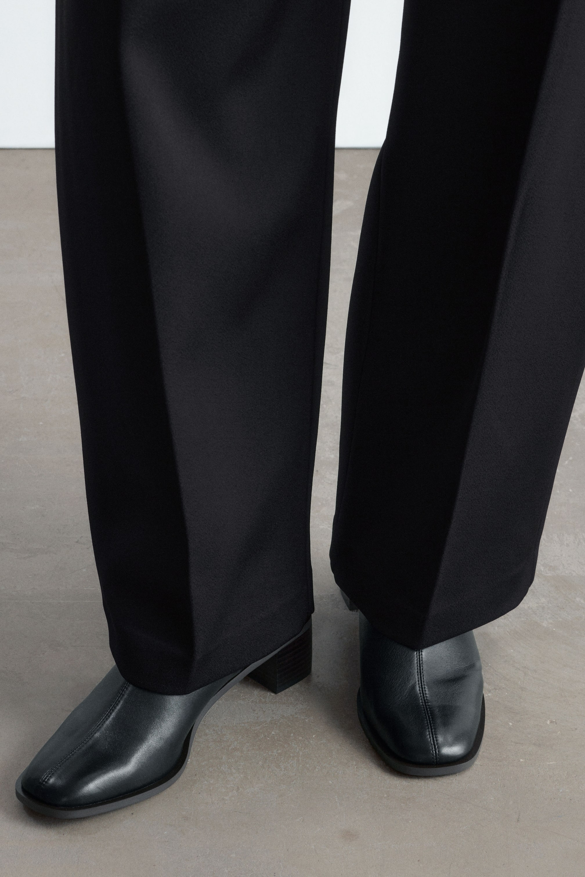 Grösseres Bild ansehen: Schwarze Sock Boots aus Nappaleder mit eckiger Spitze, mittiger Naht vorne und niedrigem Blockabsatz werden vom Model getragen. Die Stiefel sind unter weit geschnittenen schwarzen Hosen gestylt.