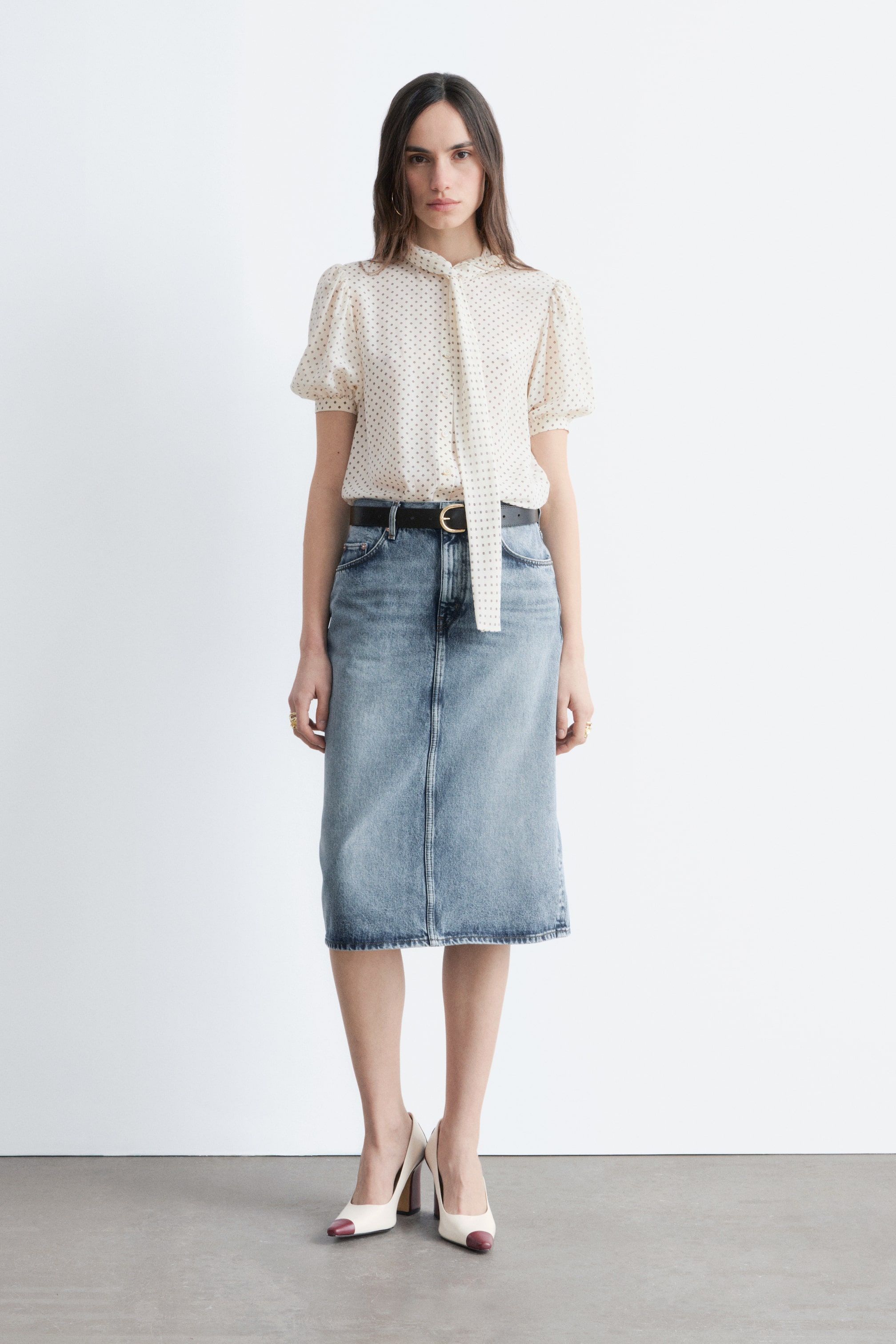 Vezi imaginea mai mare: O bluză de culoare crem, cu buline închise la culoare, mâneci scurte bufante și o cravată lungă la gât, este asortată cu o fustă midi din denim albastru deschis. O curea neagră cu o cataramă aurie subliniază talia, iar pantofii cu vârf ascuțit în două tonuri, crem și burgundy, completează ansamblul.