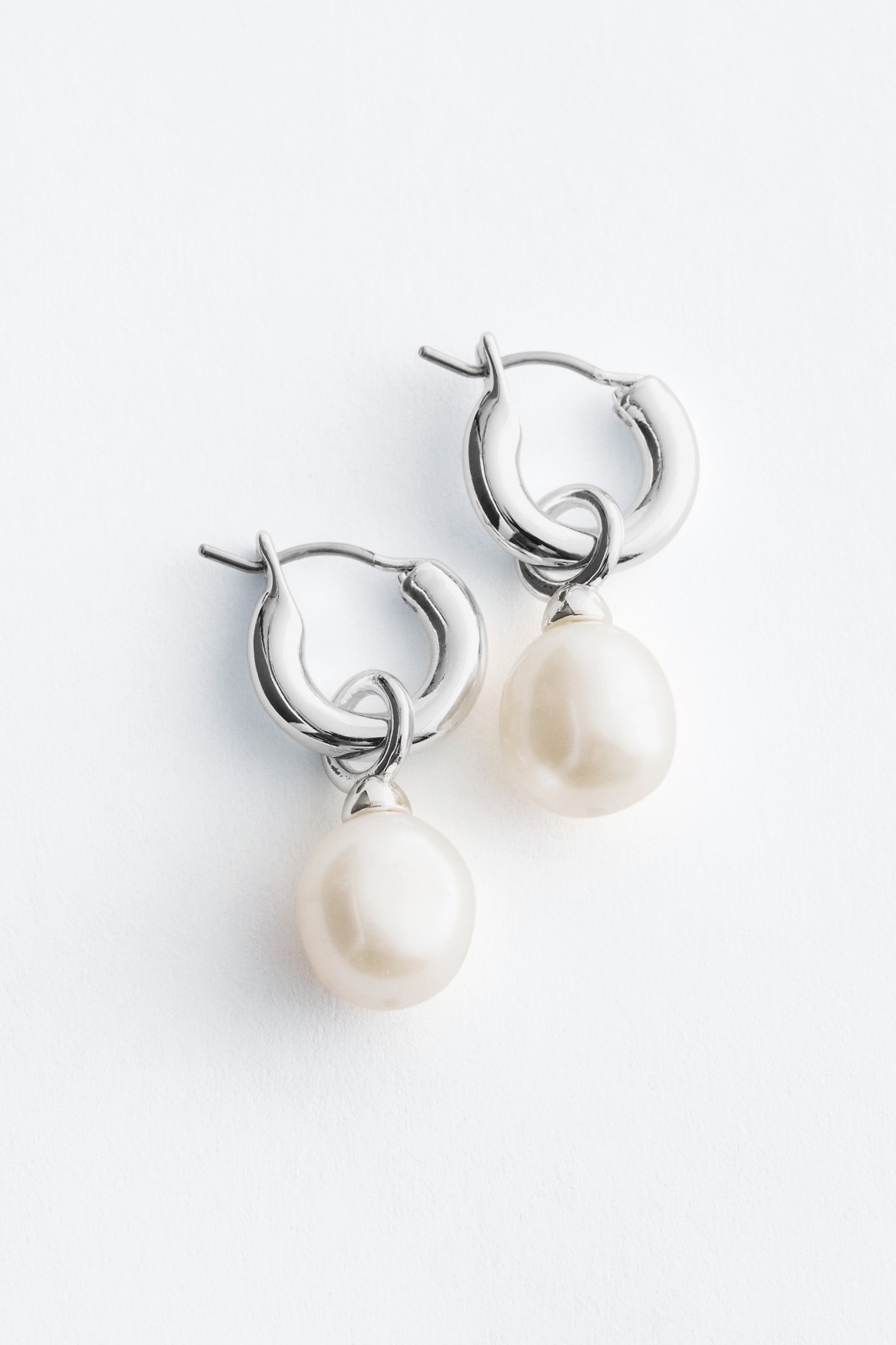 Προβολή μεγαλύτερης εικόνας: Ένα ζευγάρι σκουλαρίκια που διαθέτει δύο χοντρούς, ασημί κρίκους, καθένας από τους οποίους αναρτά μια λεία, σφαιρική, υπόλευκη πέρλα και παρουσιάζει ένα γυαλισμένο φινίρισμα.