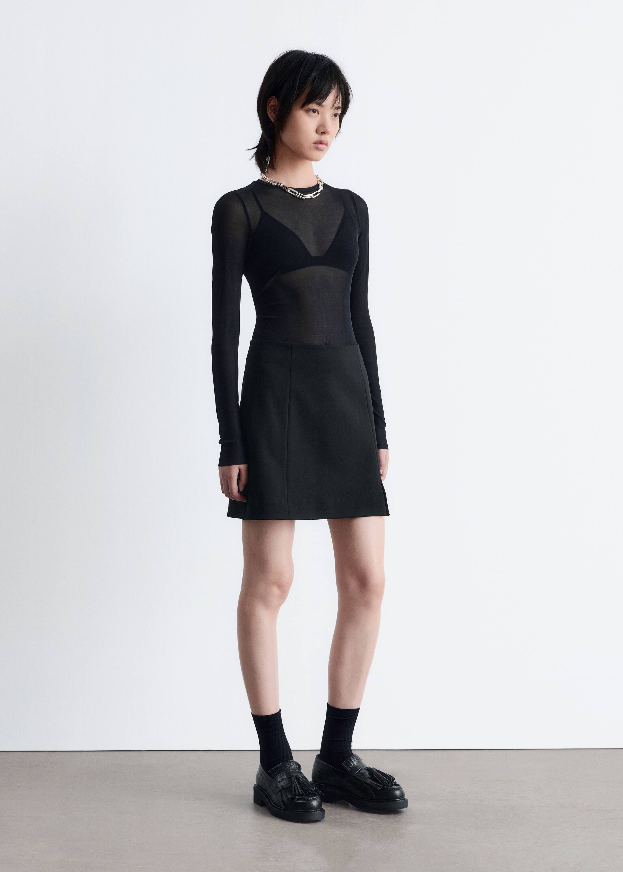 Agrandir l'image: Un haut noir transparent à manches longues est porté par-dessus un soutien-gorge noir, associé à une mini-jupe noire avec des détails de coutures verticales subtiles. L'ensemble est présenté sous un léger angle, complété par des mocassins à glands noirs avec une semelle épaisse, des chaussettes côtelées noires et un collier à maillons épais de couleur argentée.
