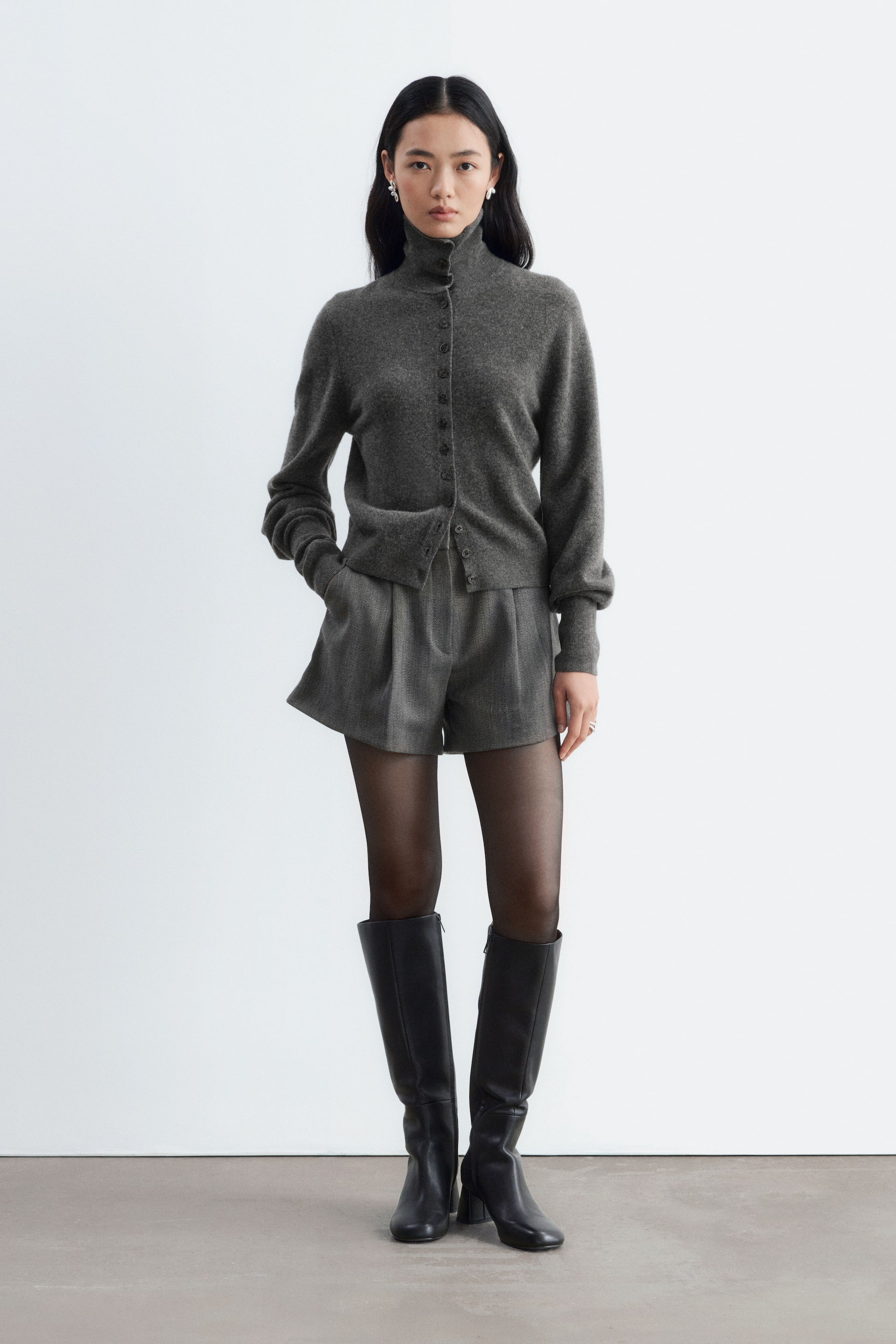 View larger image: Grey wool-blend shorts featuring front pleats and an A-line silhouette are being worn by the model, who is standing with one hand in the side pocket. The shorts are styled with a matching grey buttoned cardigan, sheer black tights, and black knee-high leather boots.