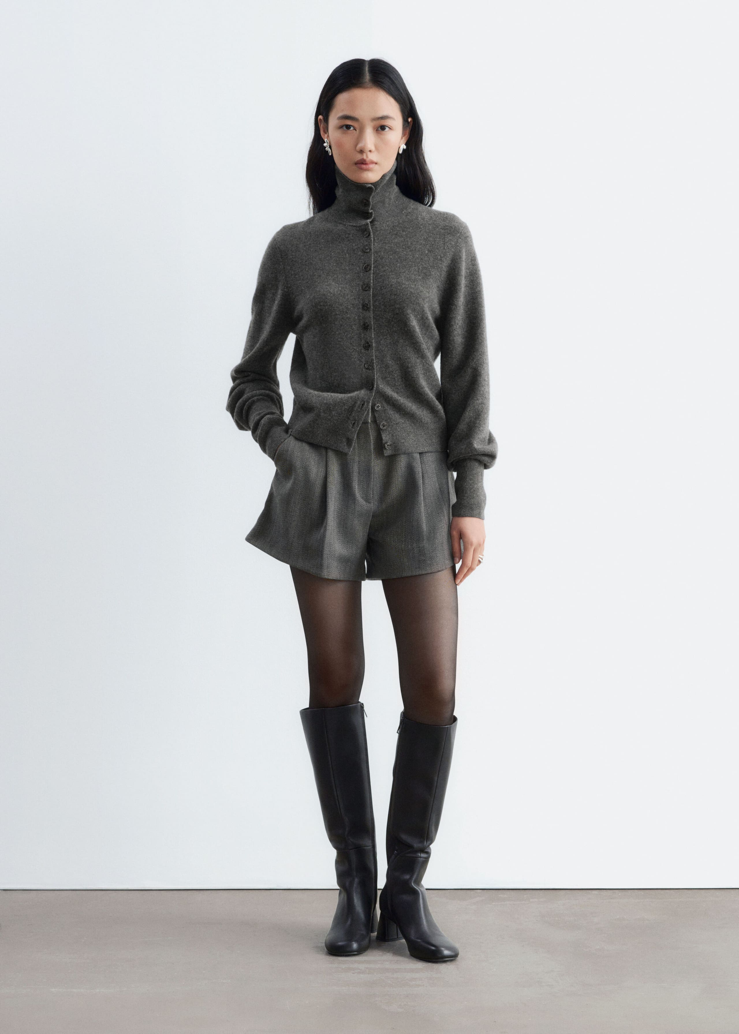 Grey wool-blend shorts featuring front pleats and an A-line silhouette are worn by the model, who is standing with one hand in the side pocket. The shorts are styled with a matching grey buttoned cardigan, sheer black tights, and black knee-high leather boots.