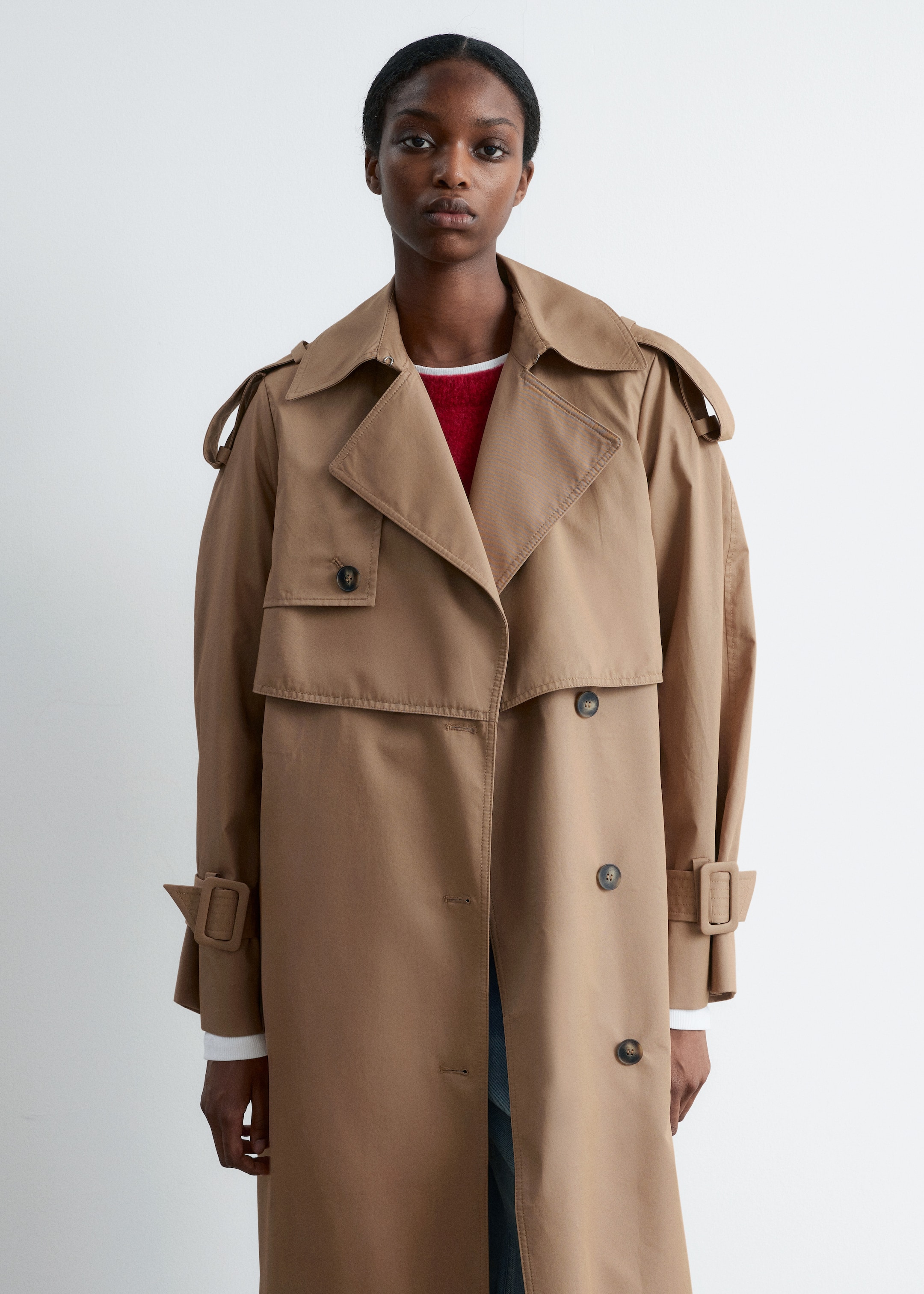 Visualizza immagine più grande: Un trench coat color cachi è indossato aperto, mettendo in mostra un davanti doppiopetto con bottoni scuri, un ampio colletto con revers a lancia, spalline, una pattina antipioggia anteriore e cinturini con fibbia ai polsini. Un maglione rosso con un colletto bianco è visibile sotto.