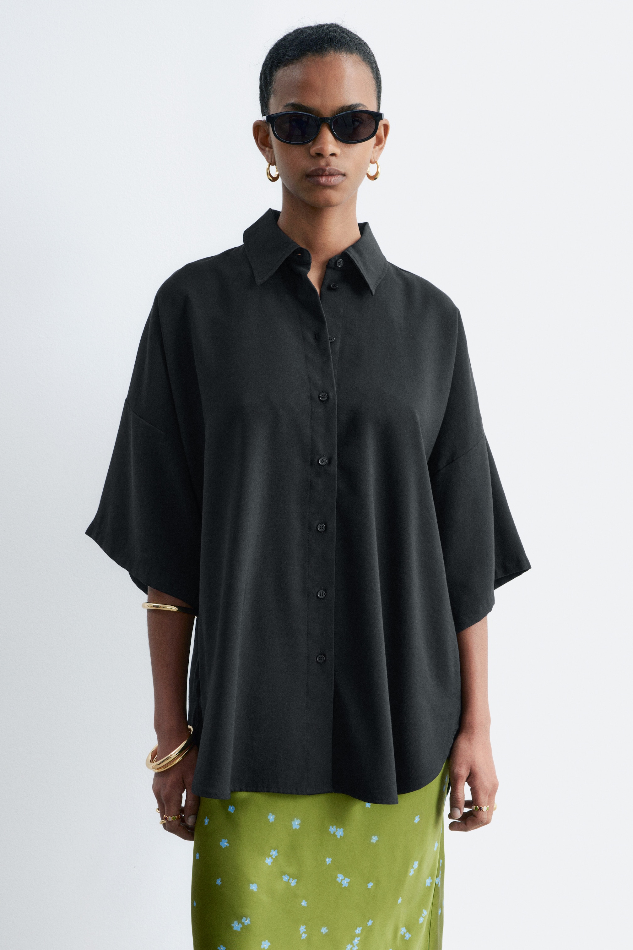 Grösseres Bild ansehen: Ein schwarzes, übergroßes Hemd mit klassischem Kragen, Knopfleiste vorne und weiten, ellbogenlangen Ärmeln wird vom Model über einem grün gemusterten Rock getragen.