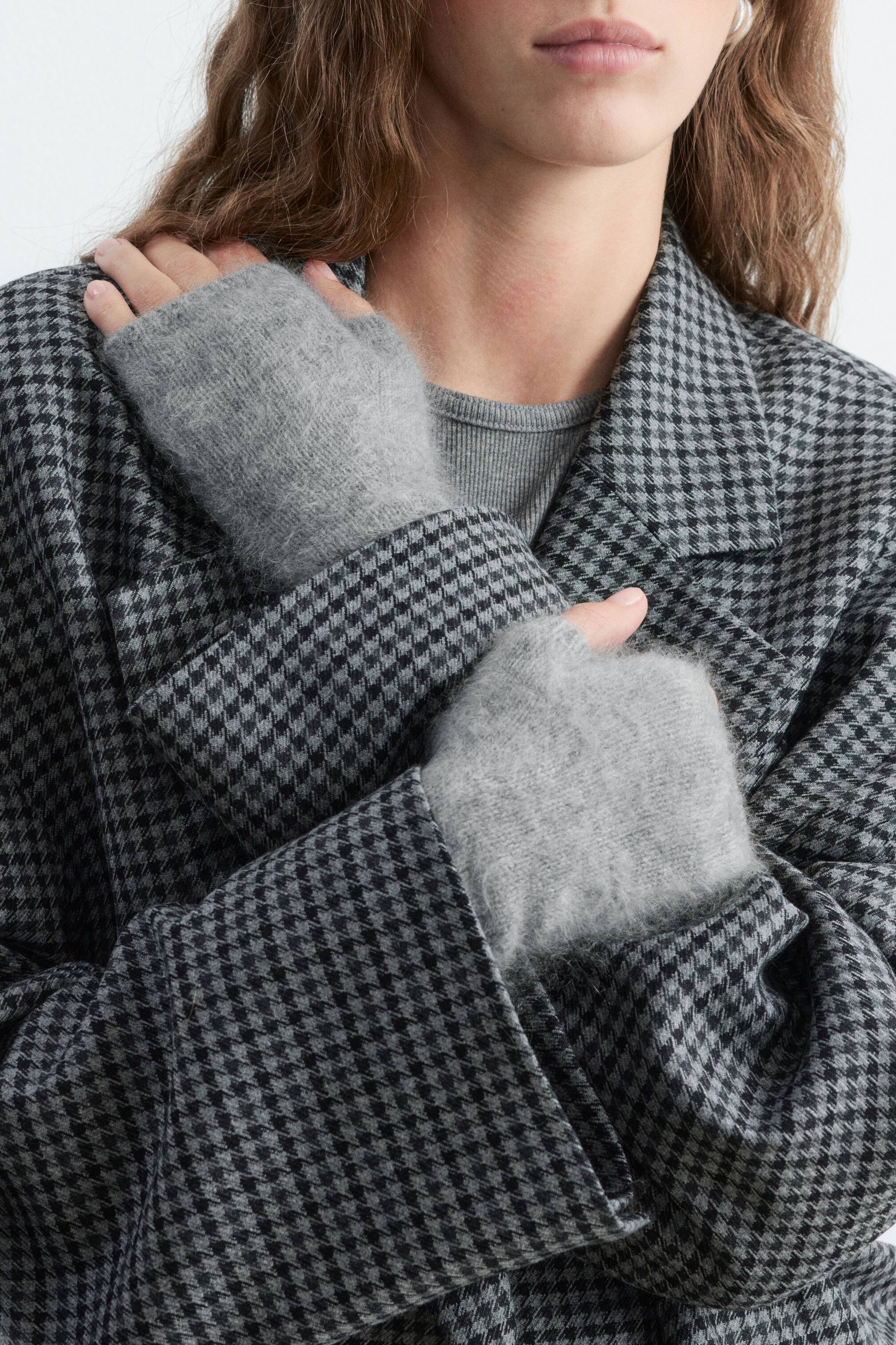 View larger image: Grey fingerless gloves crafted from brushed cashmere are being worn by the model, who is crossing their arms over a black and grey houndstooth coat. The soft, textured gloves cover the hands and wrists while leaving the fingers and thumbs exposed.