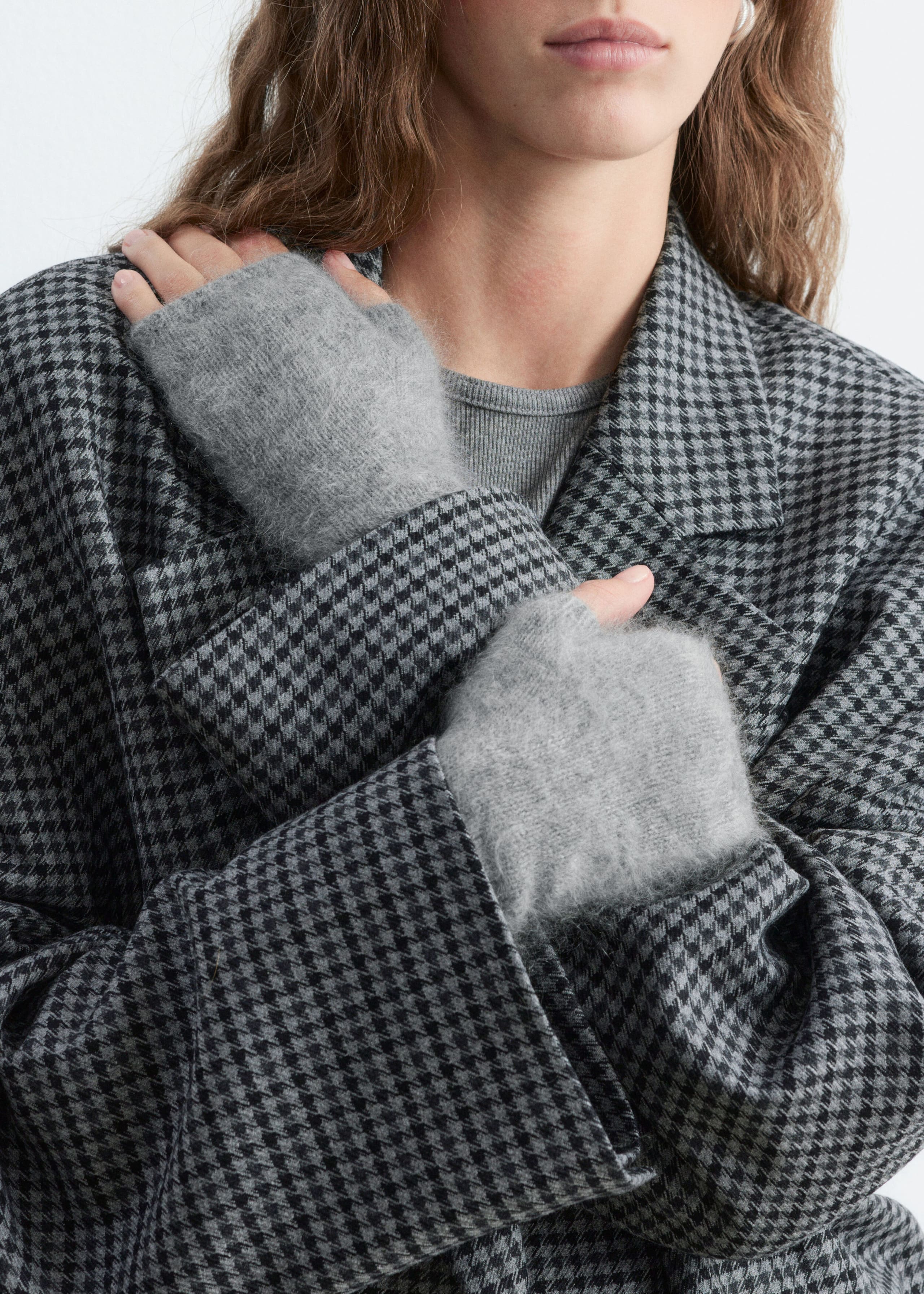 Grey fingerless gloves crafted from brushed cashmere are being worn by the model, who is crossing their arms over a black-and-gray houndstooth coat. The soft, textured gloves cover the hands and wrists while leaving the fingers and thumbs exposed.
