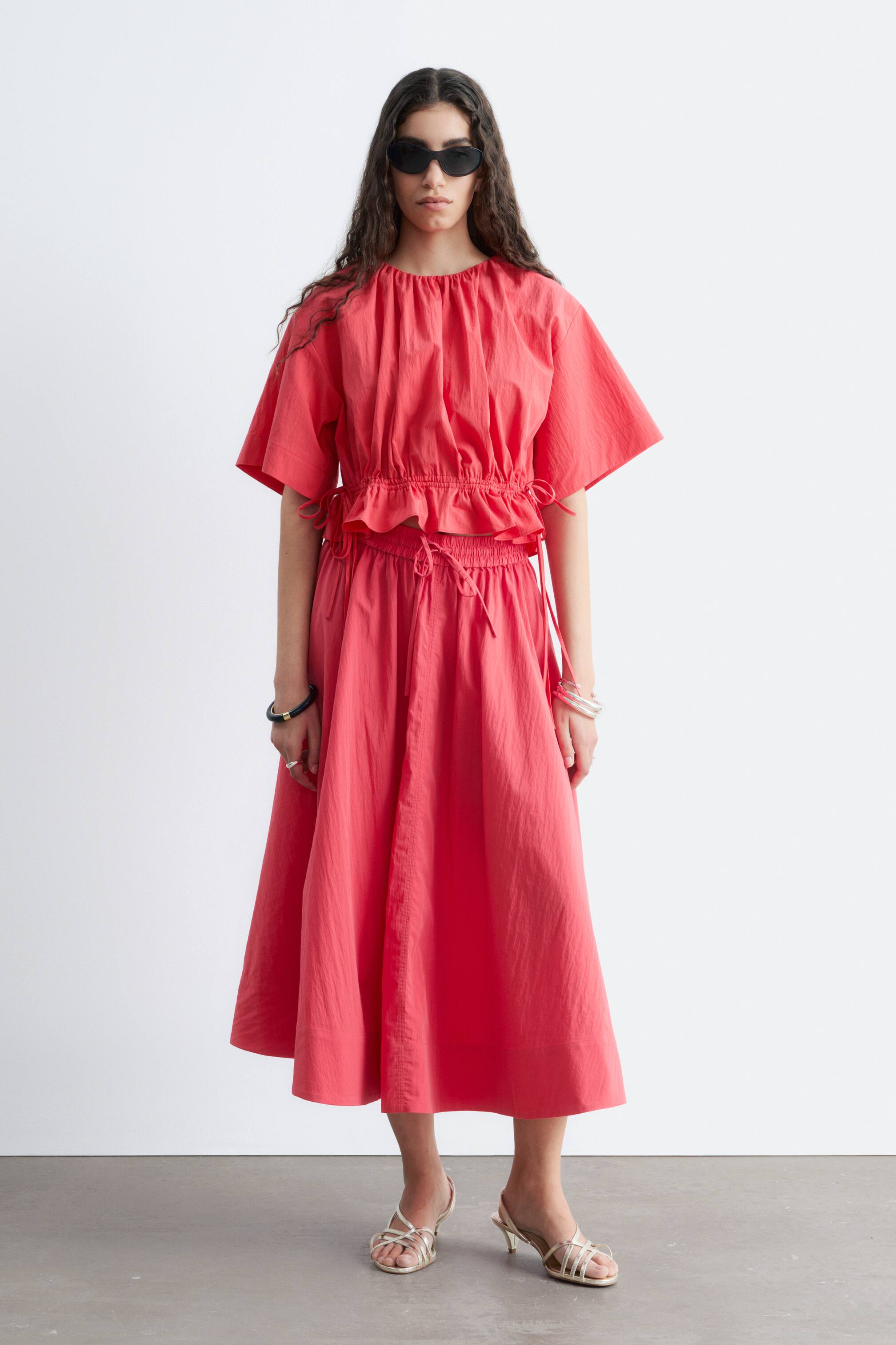 Ver imagen más grande: La modelo lleva una falda larga de color coral, que presenta una cinturilla elástica fruncida con cierre de lazo en la parte delantera. La falda tiene una silueta voluminosa y fluida y se combina con un top corto a juego y sandalias de tiras de color claro.