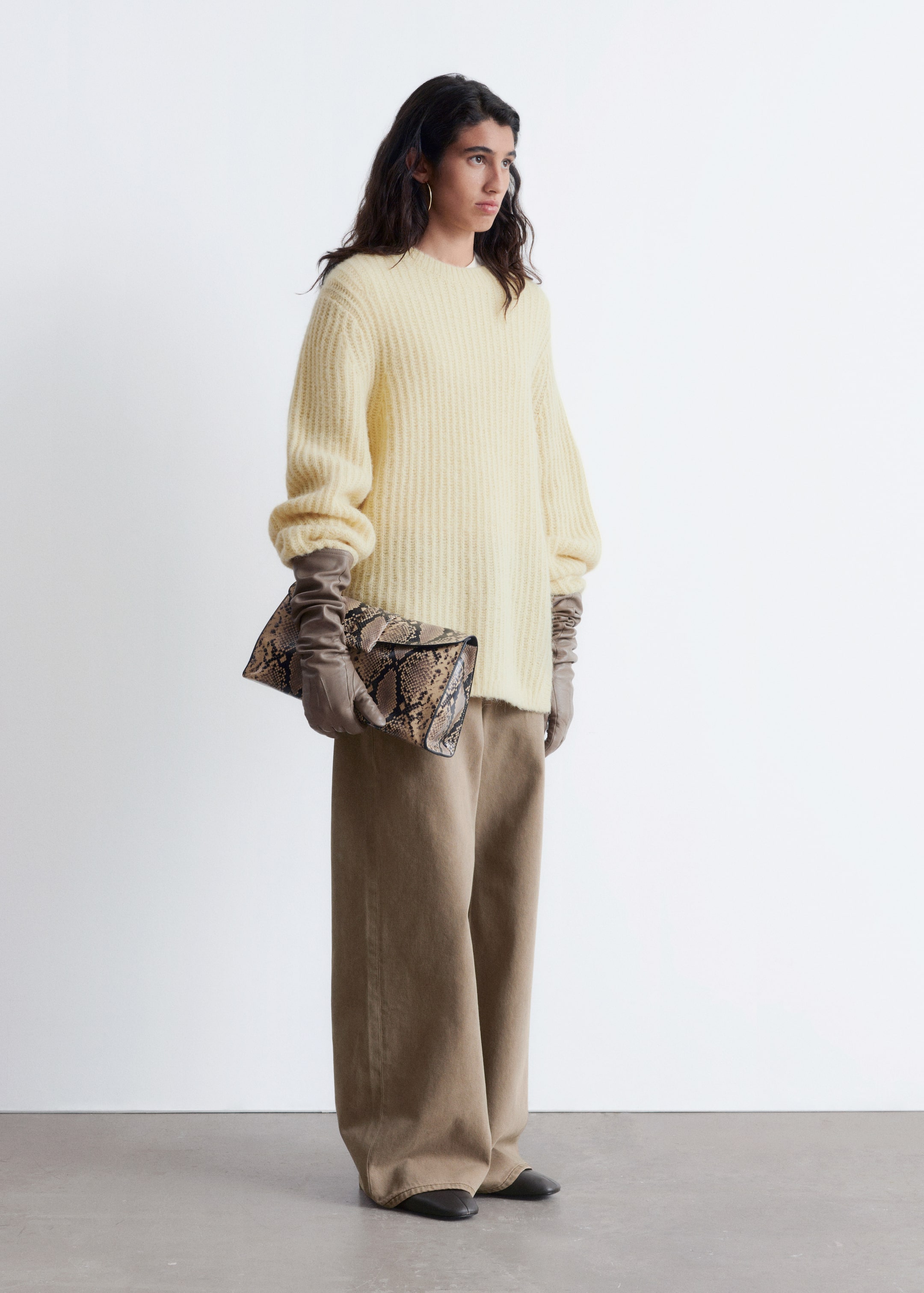 Visualizza immagine più grande: Un maglione a girocollo in maglia a coste di colore giallo chiaro, caratterizzato da una vestibilità oversize e maniche voluminose che si raccolgono ai polsi, viene mostrato da un'angolazione leggermente laterale. Viene abbinato a pantaloni a gamba larga e a tutta lunghezza di colore marrone chiaro, guanti lunghi color talpa e scarpe basse scure, mentre una pochette pieghevole con stampa pitonata marrone e nera viene tenuta in mano.