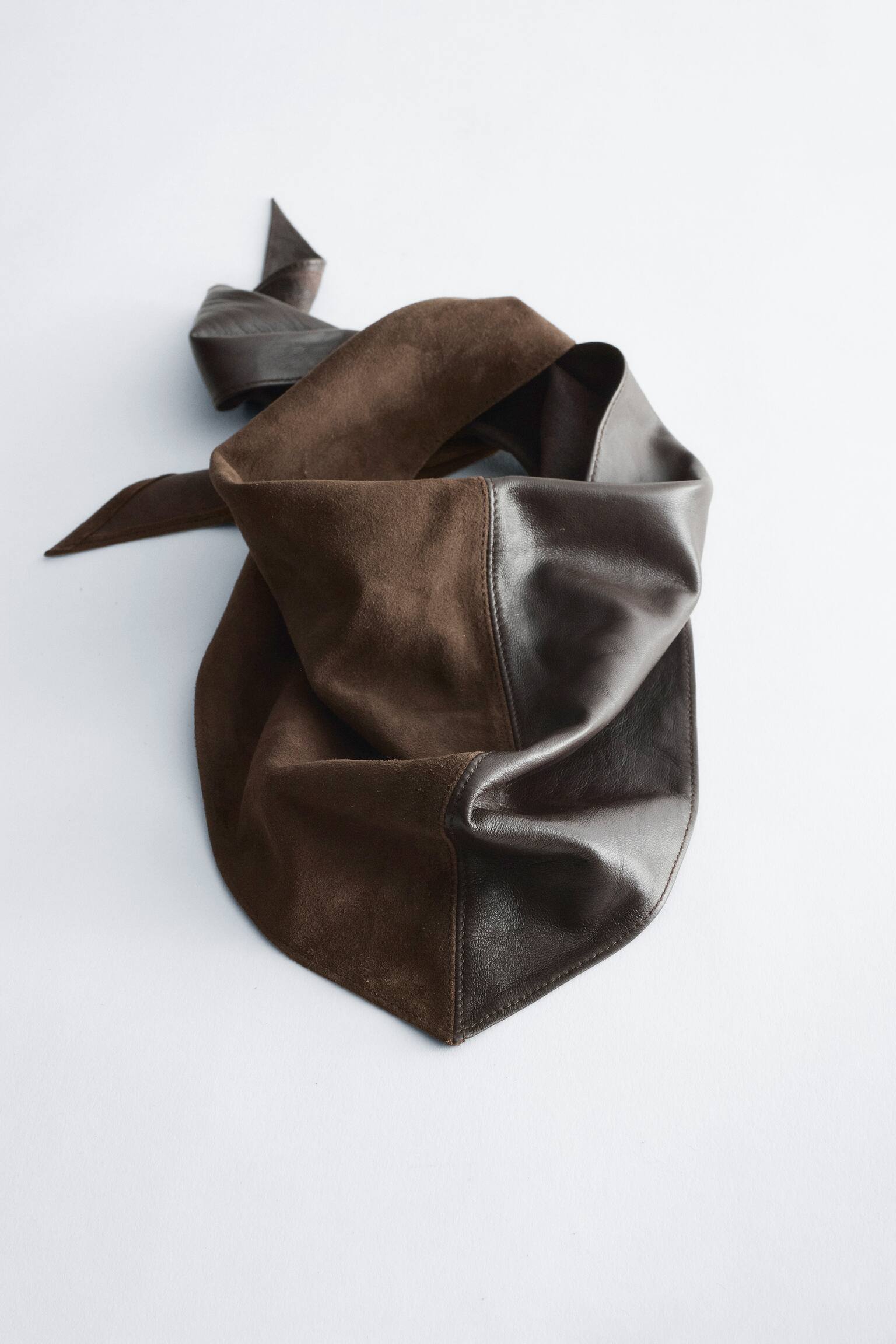 Um bandana triangular castanho-escuro apresenta, visto de frente, um design de textura dividida, com uma metade em camurÃ§a mate e a outra metade em pele lisa. O bandana repousa num arranjo drapeado que destaca a sua ponta frontal e o contraste entre as texturas.