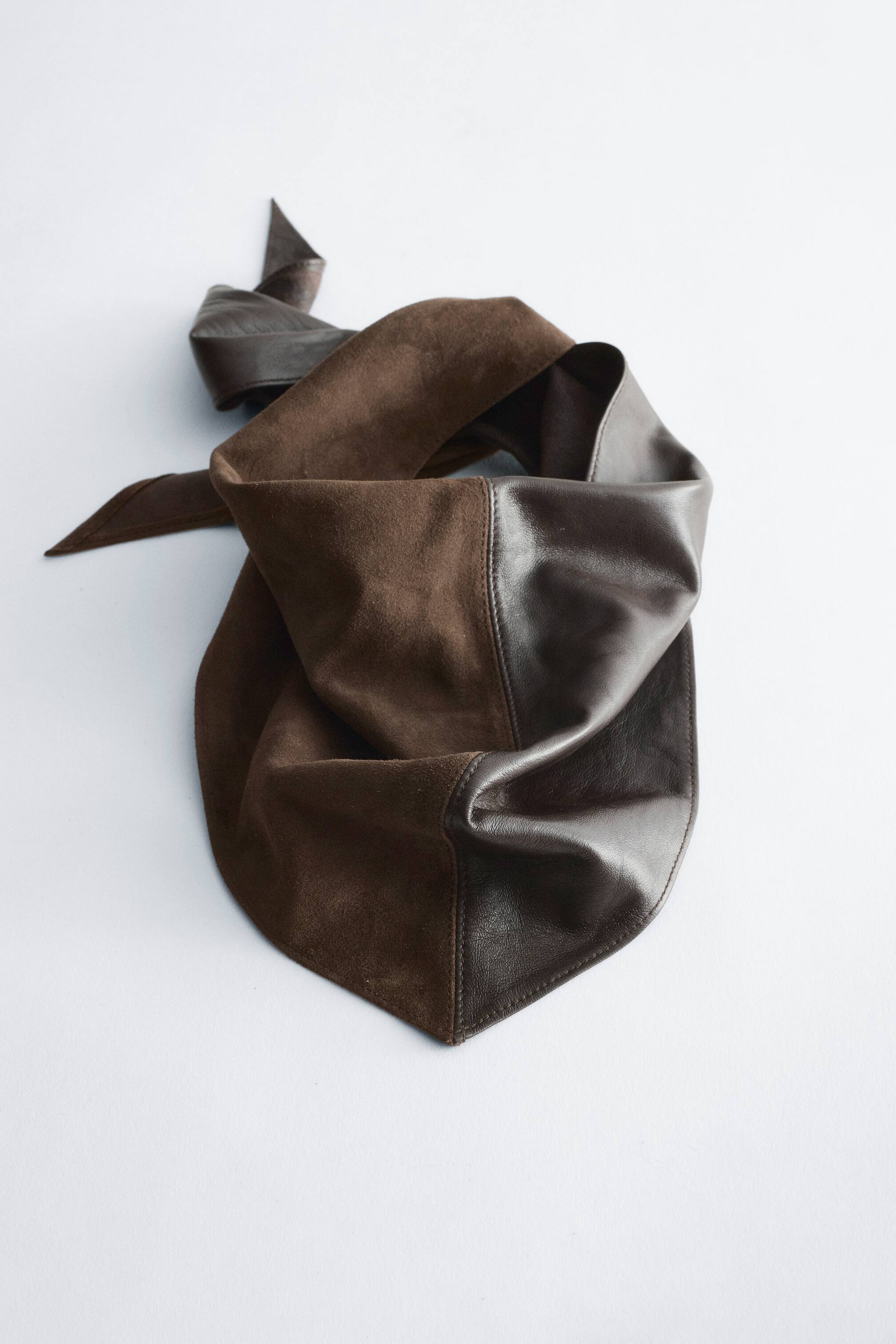 Ver imagem maior: Um bandana triangular castanho-escuro apresenta, visto de frente, um design de textura dividida, com uma metade em camurça mate e a outra metade em pele lisa. O bandana repousa num arranjo drapeado que destaca a sua ponta frontal e o contraste entre as texturas.