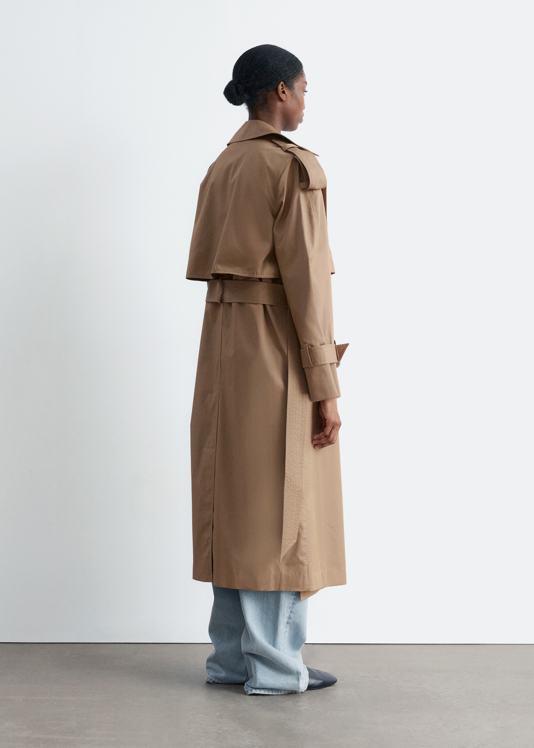 Visualizza immagine più grande: Un lungo trench coat color sabbia viene visto da dietro, con una pattina antipioggia sulla parte superiore della schiena, spalline sulle spalle e una cintura dello stesso tessuto in vita. Le cinghie regolabili sui polsini sono visibili sulle maniche, e l'orlo del cappotto si estende sotto le ginocchia, coprendo jeans a lavaggio chiaro.