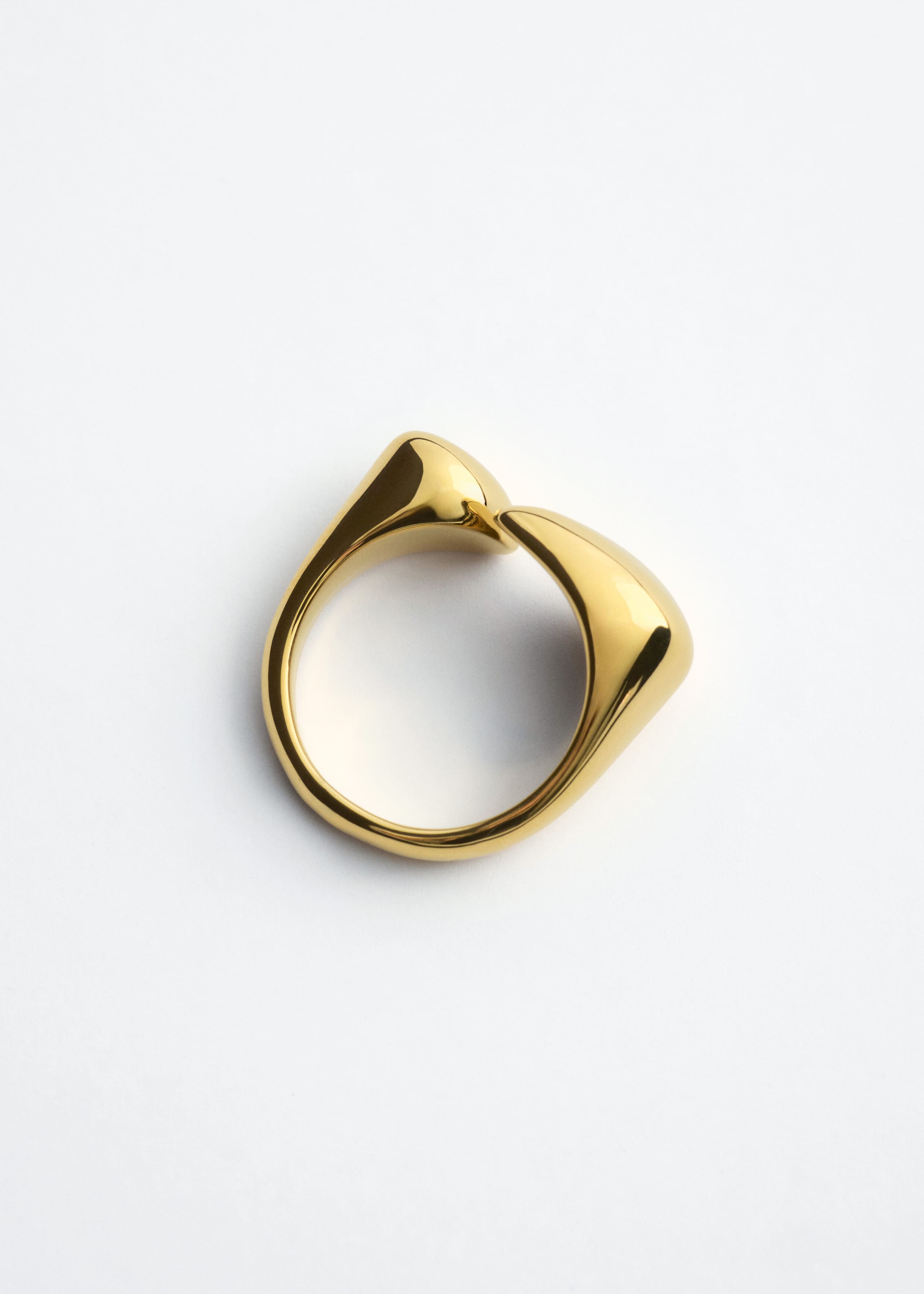Ein polierter Messingring liegt flach und präsentiert sein offenes Banddesign mit zwei asymmetrischen, abgerundet-skulpturalen Enden. Der Ring zeigt eine hochglänzende, reflektierende Oberfläche entlang seiner fließenden, organischen Form.