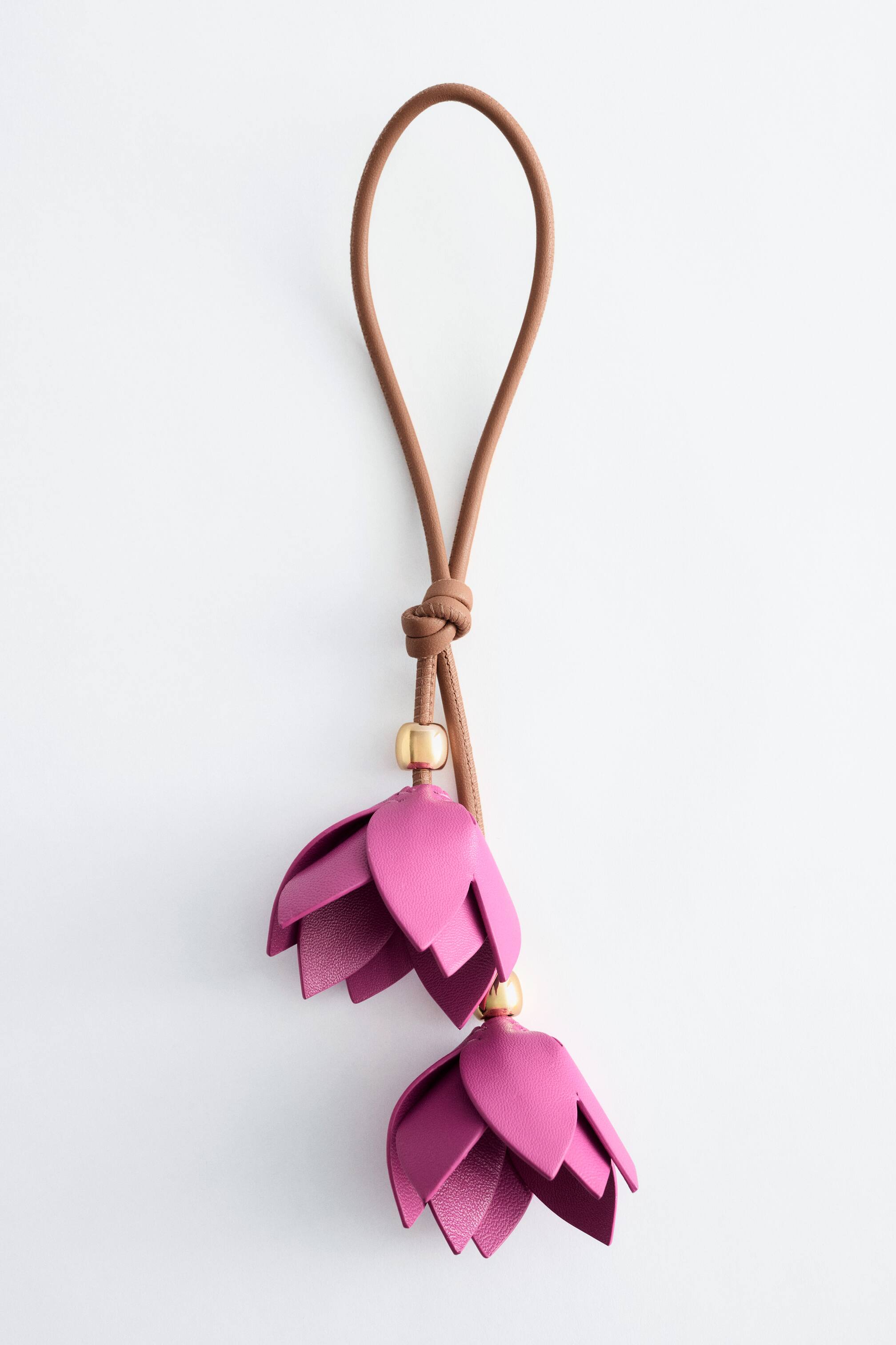 Agrandir l'image: Un charm de sac en forme de fleur présente une sangle marron en boucle, nouée à sa base, d'où pendent deux éléments en forme de fleur fuchsia. Chaque fleur est construite avec plusieurs pétales superposés et est attachée à la sangle via une petite perle dorée.