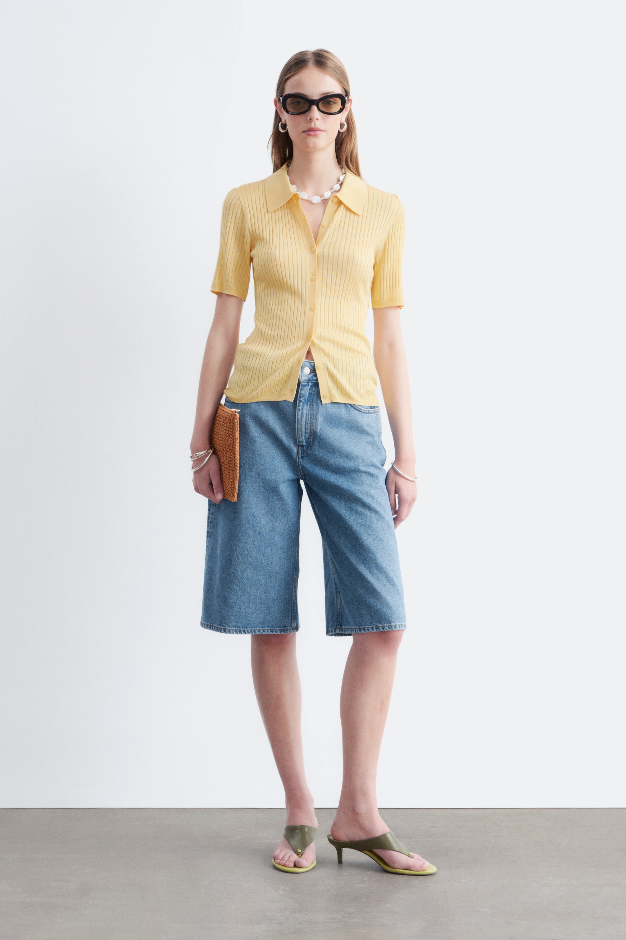 Agrandir l'image: Un polo en maille côtelée jaune à manches courtes est présenté, avec un col pointu et une fermeture boutonnée sur le devant avec des boutons de couleur claire. Sa silhouette ajustée apparaît rentrée dans un vêtement taille haute, avec les deux premiers boutons laissés défaits.
