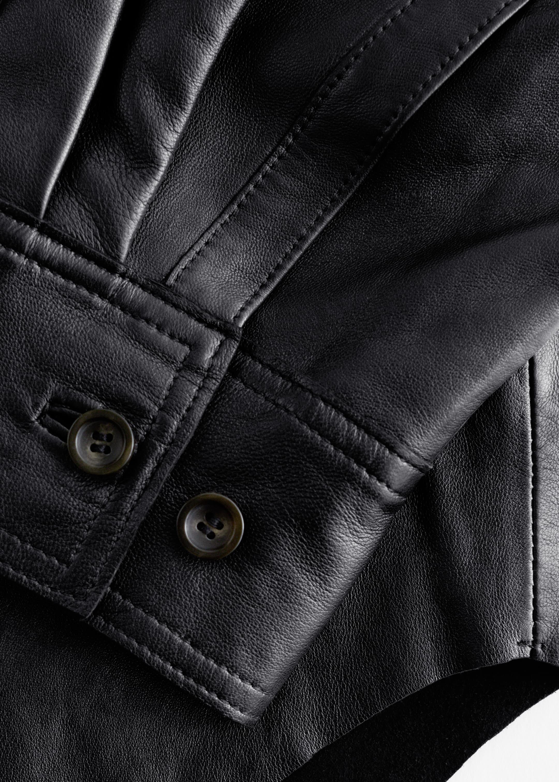 Visualizza immagine più grande: Una camicia in pelle nappa nera viene mostrata in primo piano, inclinata verso destra. Il polsino della manica presenta due bottoni scuri a quattro fori e cuciture visibili, mentre il corpo in pelle morbida mostra pieghe verticali sulla parte superiore sinistra.