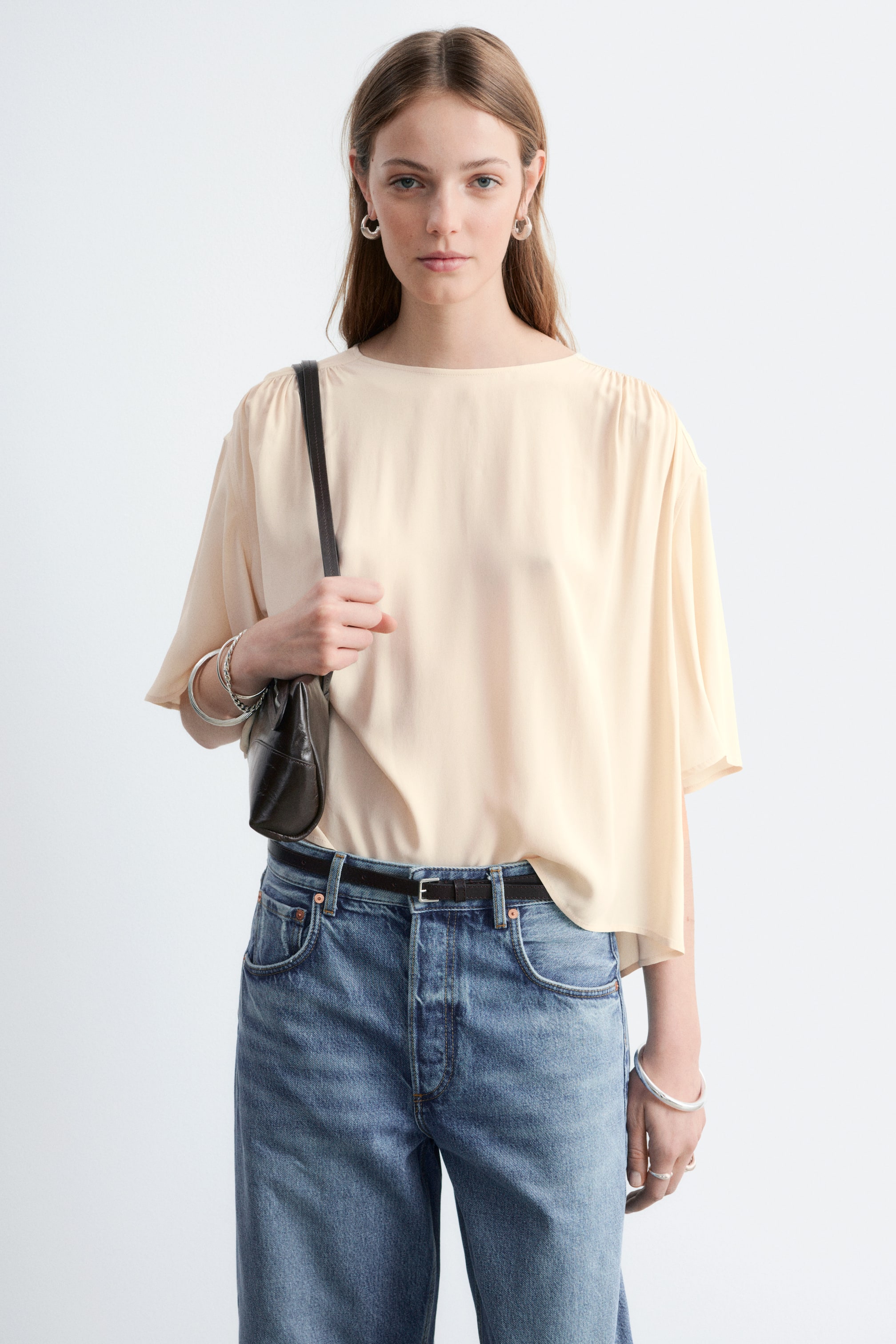 Ver imagem maior: Um top amarelo pálido está a ser apresentado, voltado para a frente. Apresenta uma gola barco, detalhes franzidos nos ombros e mangas curtas largas e fluídas. O top é desenhado com um corte descontraído e solto, e uma bainha alongada e fluída nas laterais.