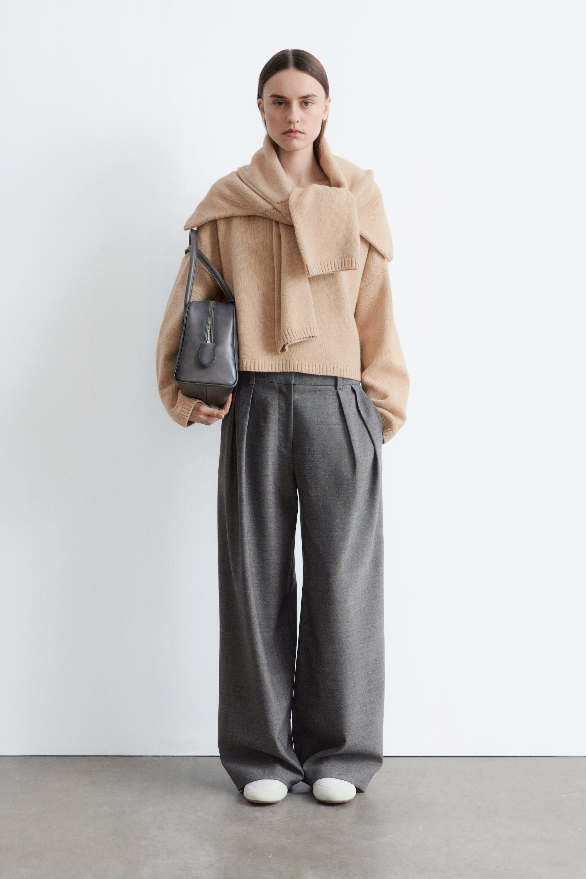 Se større billede: Et frontalt fuldkropsfoto viser en beige cropped striktrøje med lange ærmer og ribbet kant, og et matchende beige striktørklæde er draperet over skuldrene. Grå vide bukser med presninger foran fuldender underdelen, mens en struktureret grå håndtaske med top-lynlås holdes i hånden, og hvide flade sko er synlige.