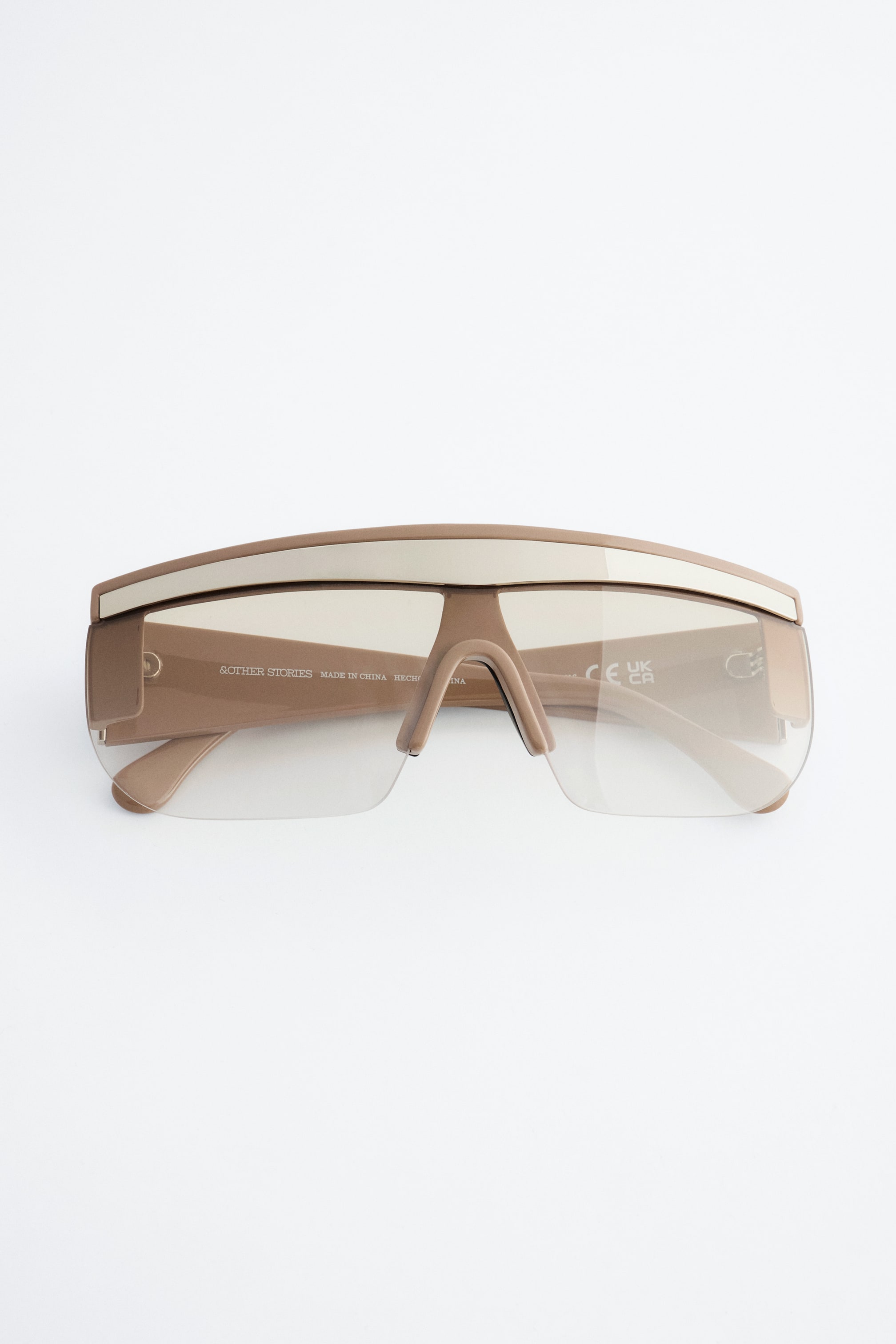 Ver imagem maior: Estes óculos de sol estilo visor estão virados para a frente, apresentando uma armação bege mate com uma faixa contrastante branca-quebrada a atravessar a barra superior. Incluem lentes semi-transparentes, ligeiramente fumadas, com a extremidade inferior sem aro e hastes bege a combinar.