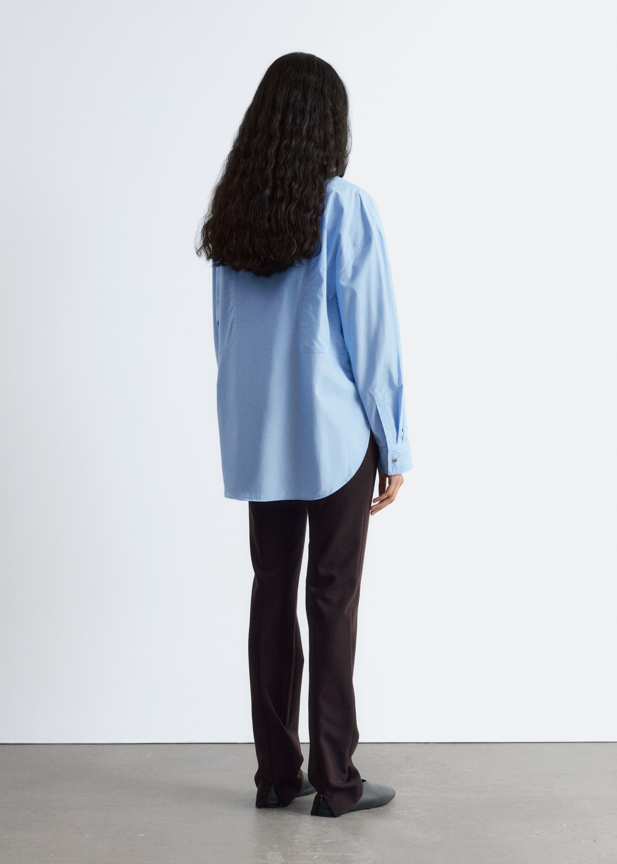 Visualizza immagine più grande: Una camicia azzurra oversize con bottoni è vista da dietro, mostrando una vestibilità rilassata, un giogo posteriore visibile, maniche lunghe con polsini abbottonati e un orlo arrotondato. È abbinata a pantaloni marrone scuro a gamba dritta con una cucitura posteriore e piccoli spacchi alla caviglia.