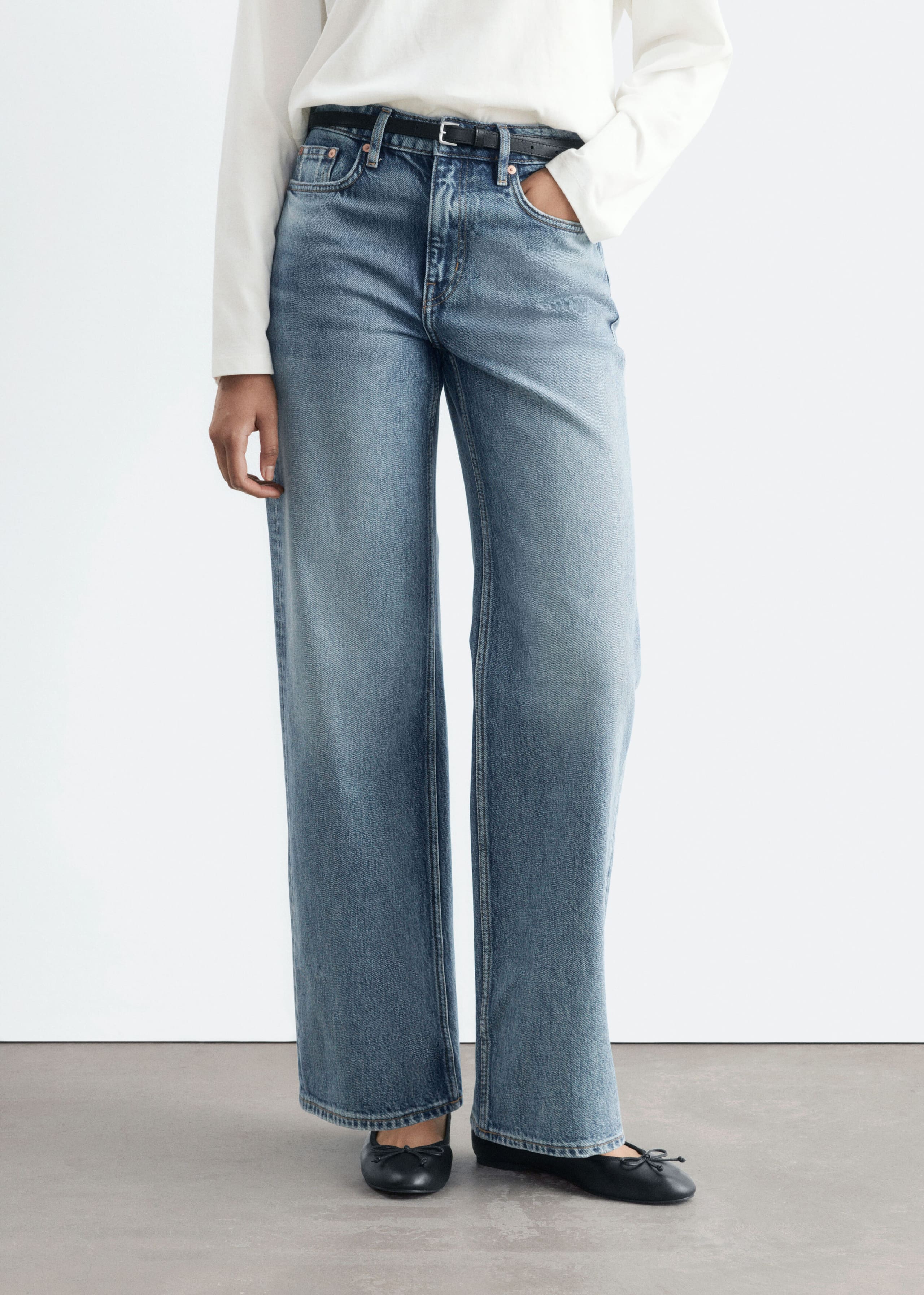 These mid-rise, straight-leg blue jeans are being worn by the model, who is standing with one hand resting in a pocket. The faded denim jeans are styled with a tucked-in white long-sleeve top, a slim black belt, and black ballet pumps.