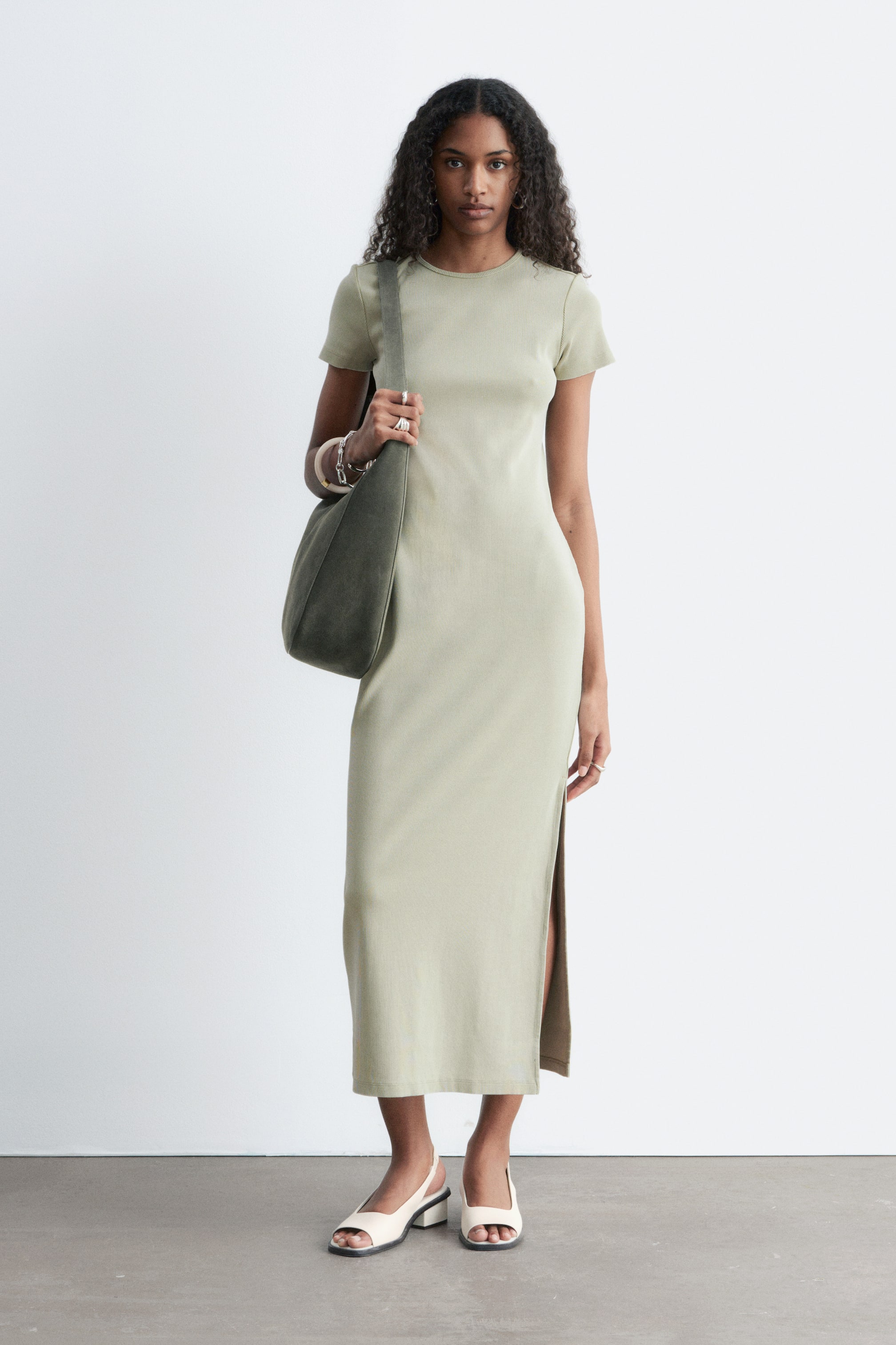 Grotere afbeelding bekijken: Een lichtgroene midijurk wordt gedragen door het model, met korte mouwen, een ronde hals en een hoge zijsplit die de nauwsluitende pasvorm benadrukt.