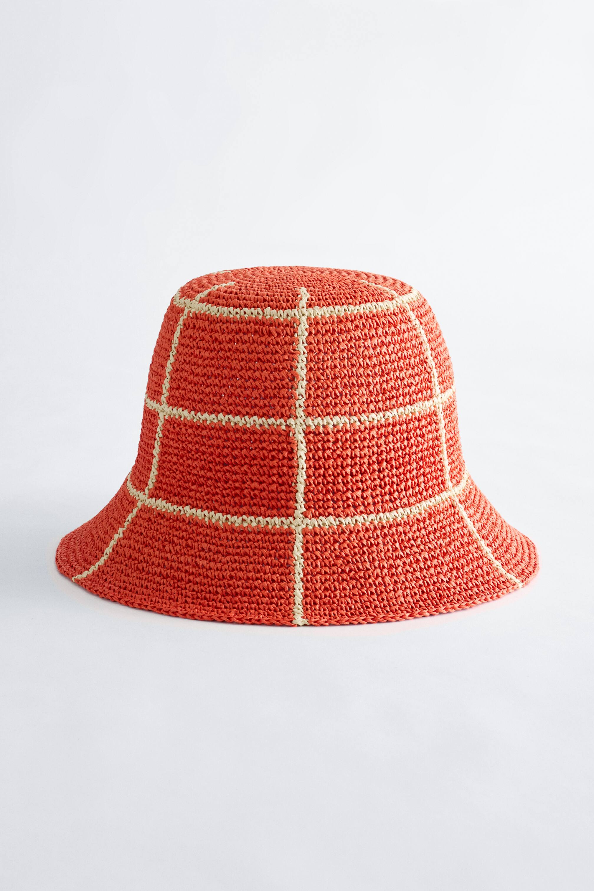 Visualizza l'immagine ingrandita: Un cappello a secchiello in paglia intrecciata arancione con un motivo a quadri di colore chiaro è leggermente rivolto in avanti, mostrando una corona strutturata e una tesa inclinata verso il basso.